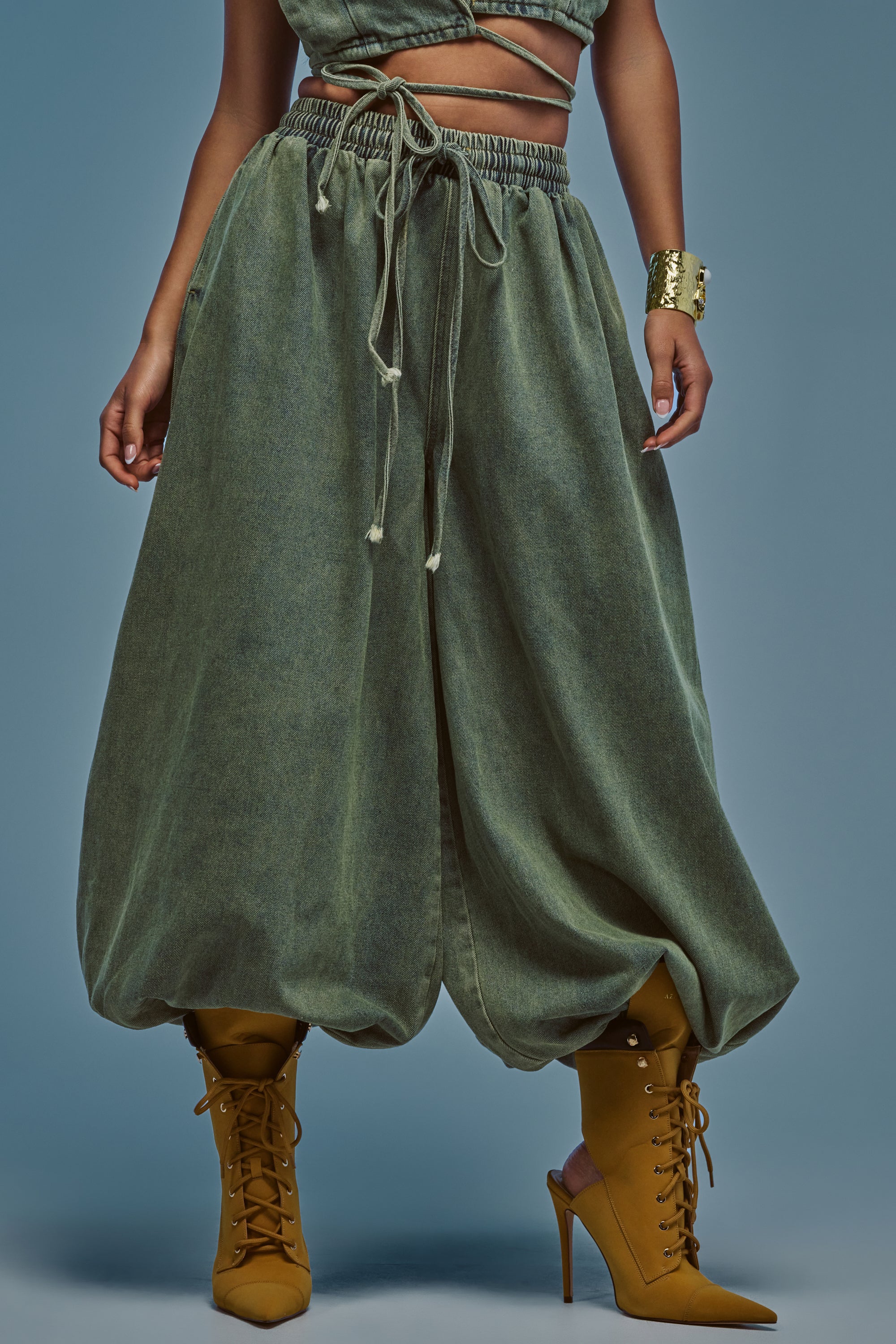 A person wearing the GUESS WHO BALLOON PANT IN DENIM, styled with a matching crop top, a gold cuff bracelet, and mustard yellow lace-up heeled boots, standing against a blue-gray background.