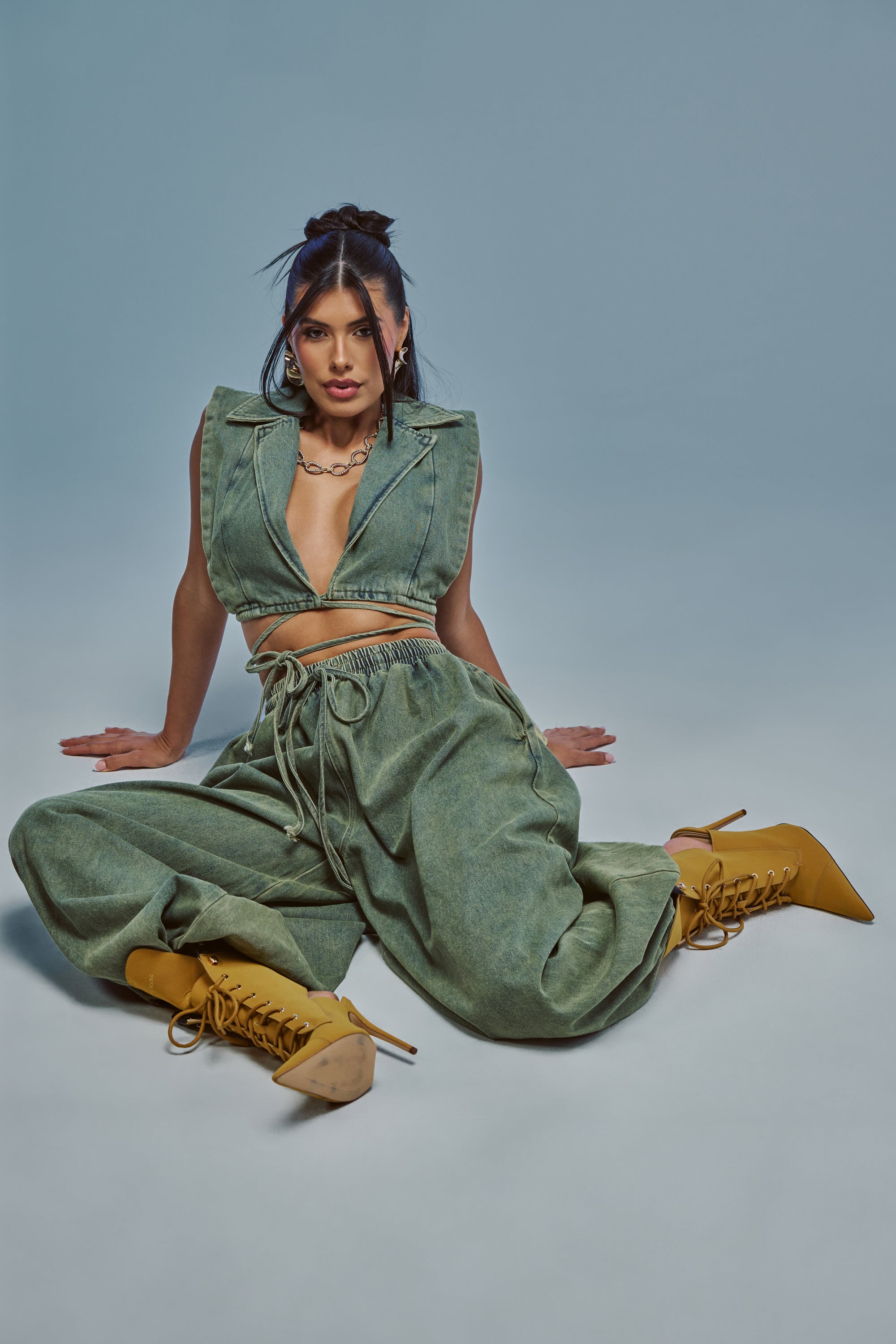 A woman sits on the floor against a plain background, wearing the GUESS WHO BALLOON PANT IN DENIM, styled with a sleeveless green denim vest, a chain necklace, and tan heeled boots. She gazes confidently at the camera, embodying effortless streetwear style.