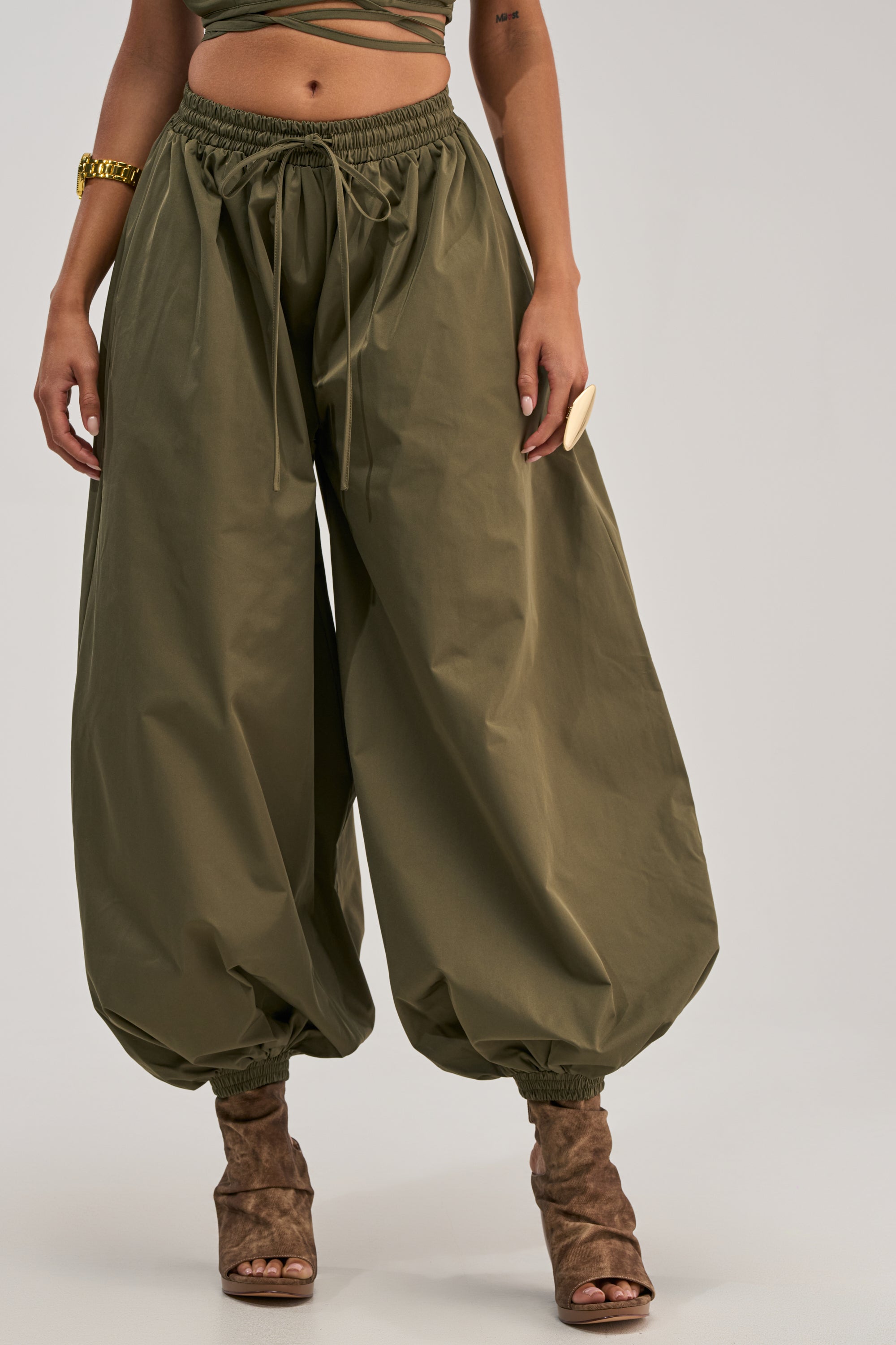 Wearing the GUESS WHO BALLOON PANT IN OLIVE, they pair its relaxed, high-rise fit and drawstring waist with brown open-toe heels, a green crop top, and gold jewelry for an effortless utility-chic look.