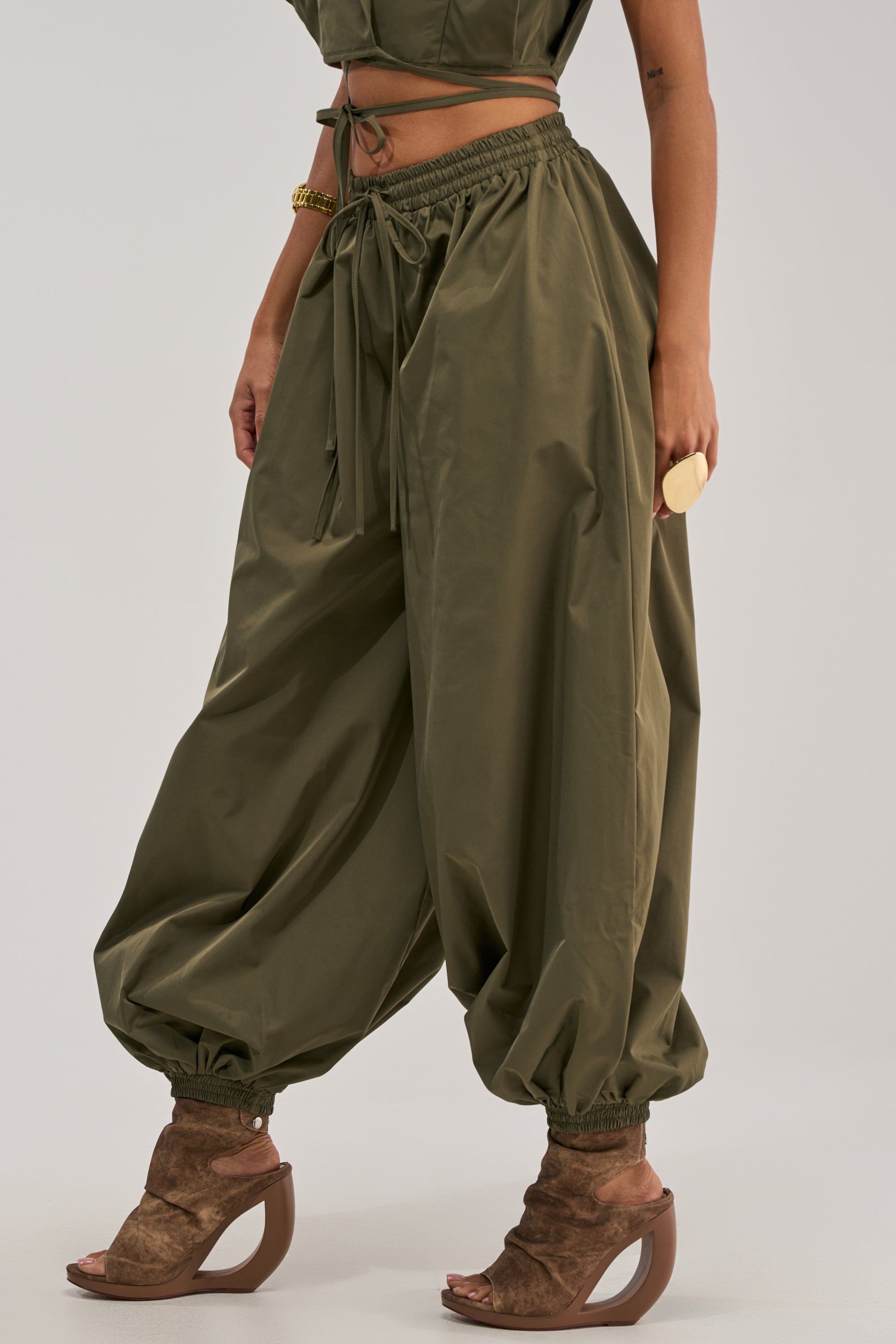 The lower half of a person is shown wearing GUESS WHO BALLOON PANT IN OLIVE with a relaxed high-rise fit and elastic ankles, styled with brown open-toe wedge heels for a utility-chic look against a plain background.