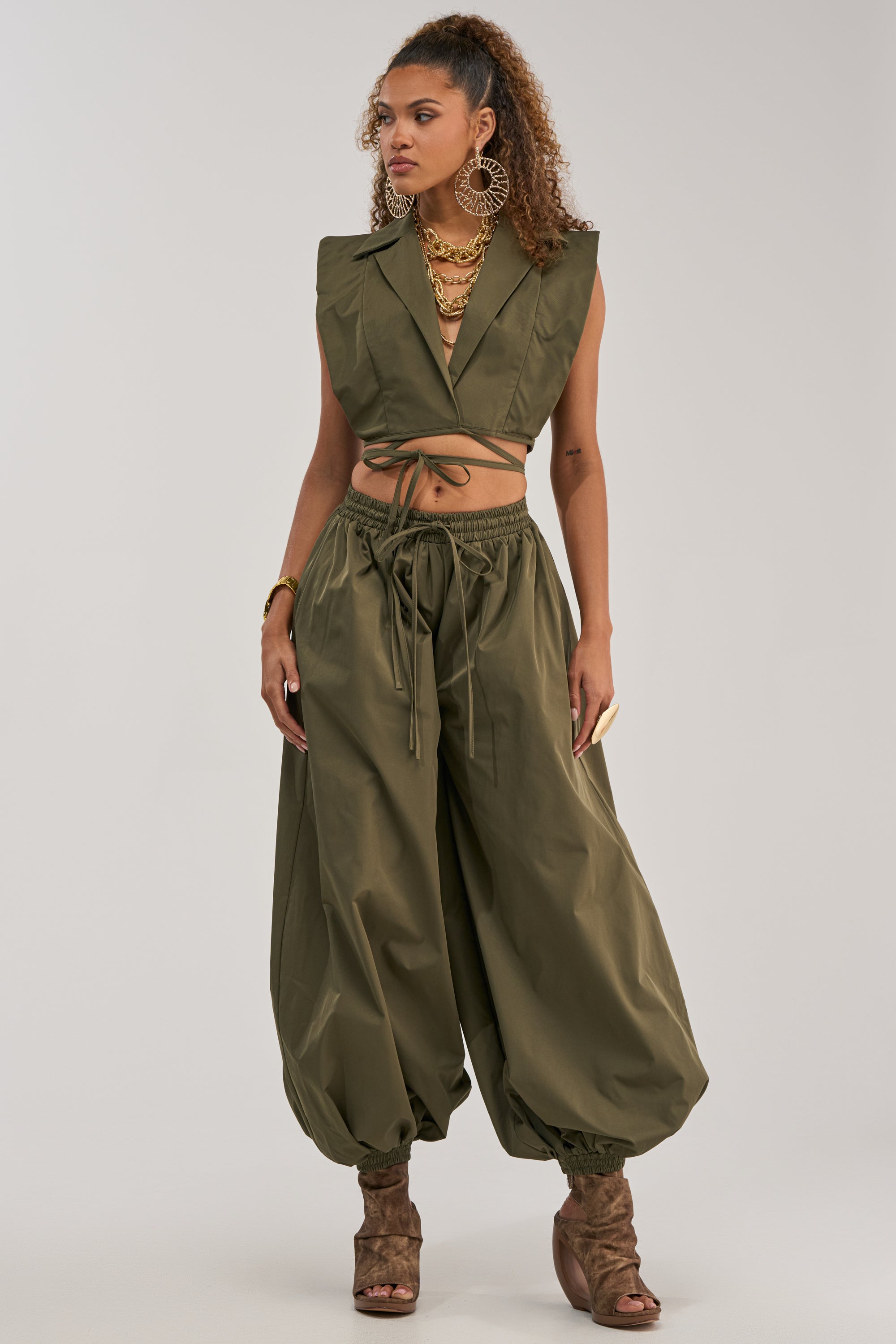 A woman stands against a plain background, effortlessly channeling utility-chic style in the GUESS WHO BALLOON PANT IN OLIVE, paired with a matching olive green sleeveless crop top, brown open-toe heels, large hoop earrings, and layered necklaces.