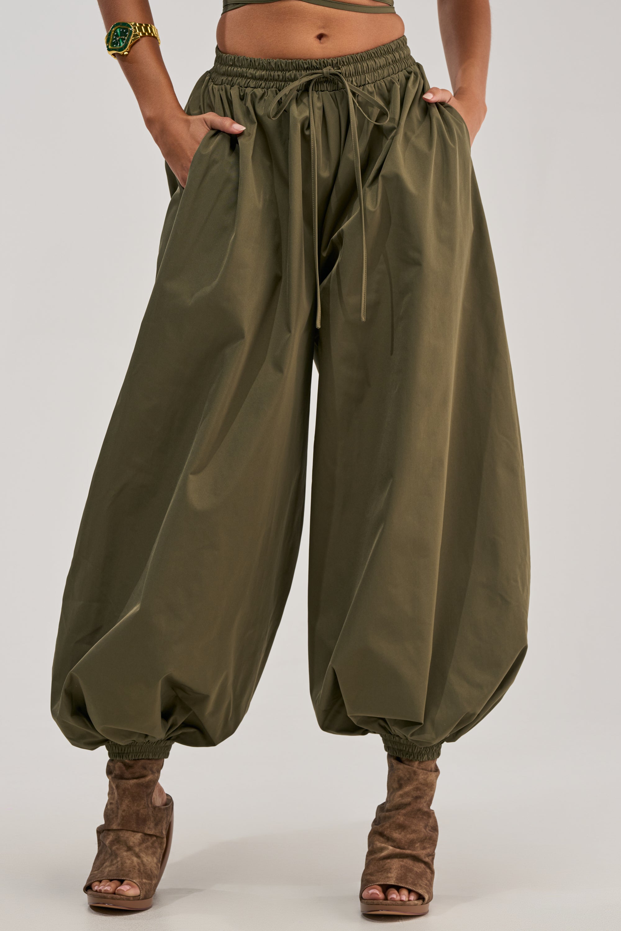 A person is wearing the GUESS WHO BALLOON PANT IN OLIVE, featuring a loose, high-rise silhouette with a drawstring waist and gathered ankles for a utility-chic look. The pants are styled with brown open-toe heeled boots and a green bracelet, hands in pockets, against a plain light-colored background.