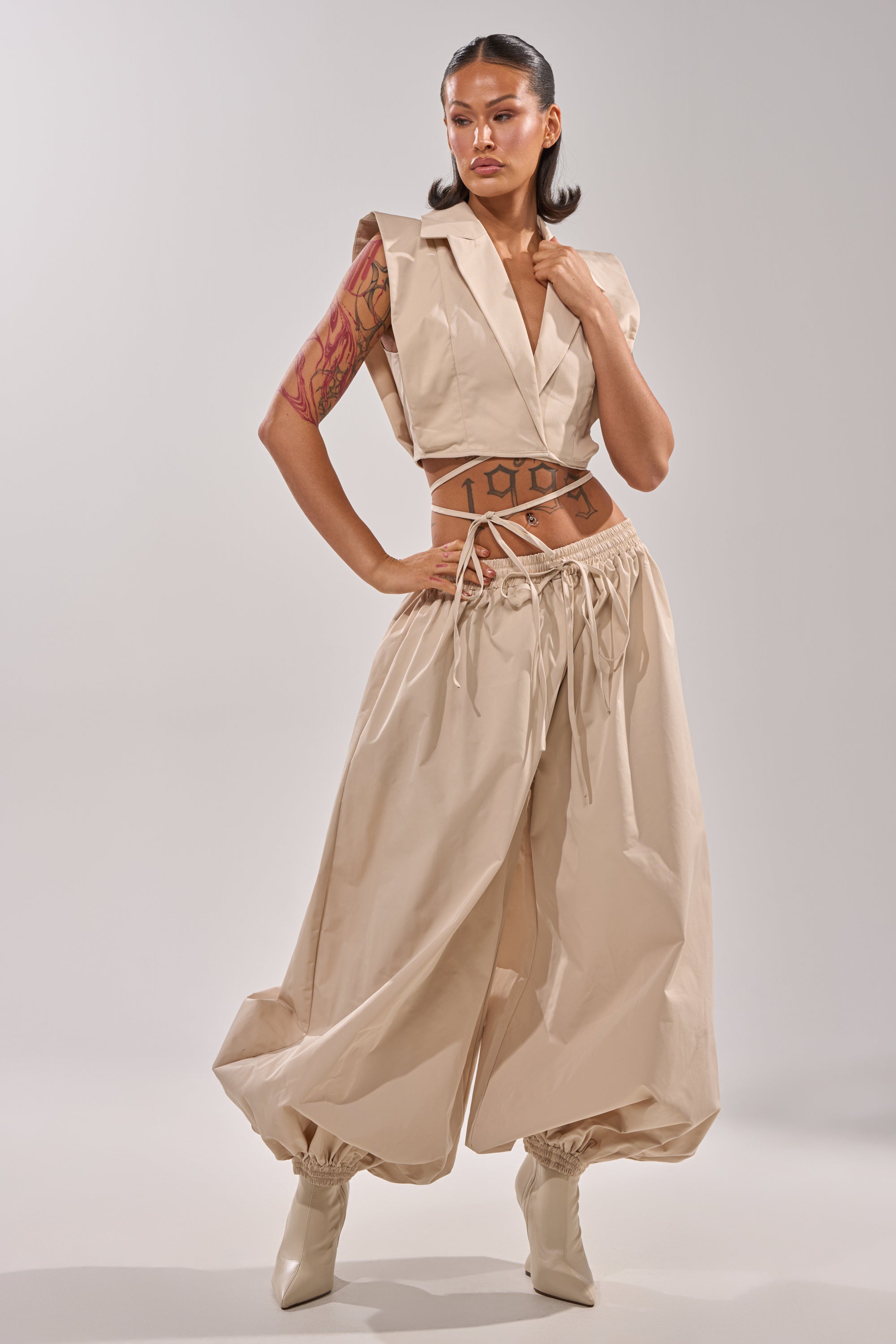 A woman with tattoos models the GUESS WHO BALLOON PANT styled with a beige sleeveless crop top, drawstring details, and light heeled boots. Her sleek hair is styled back for a confident look. 0