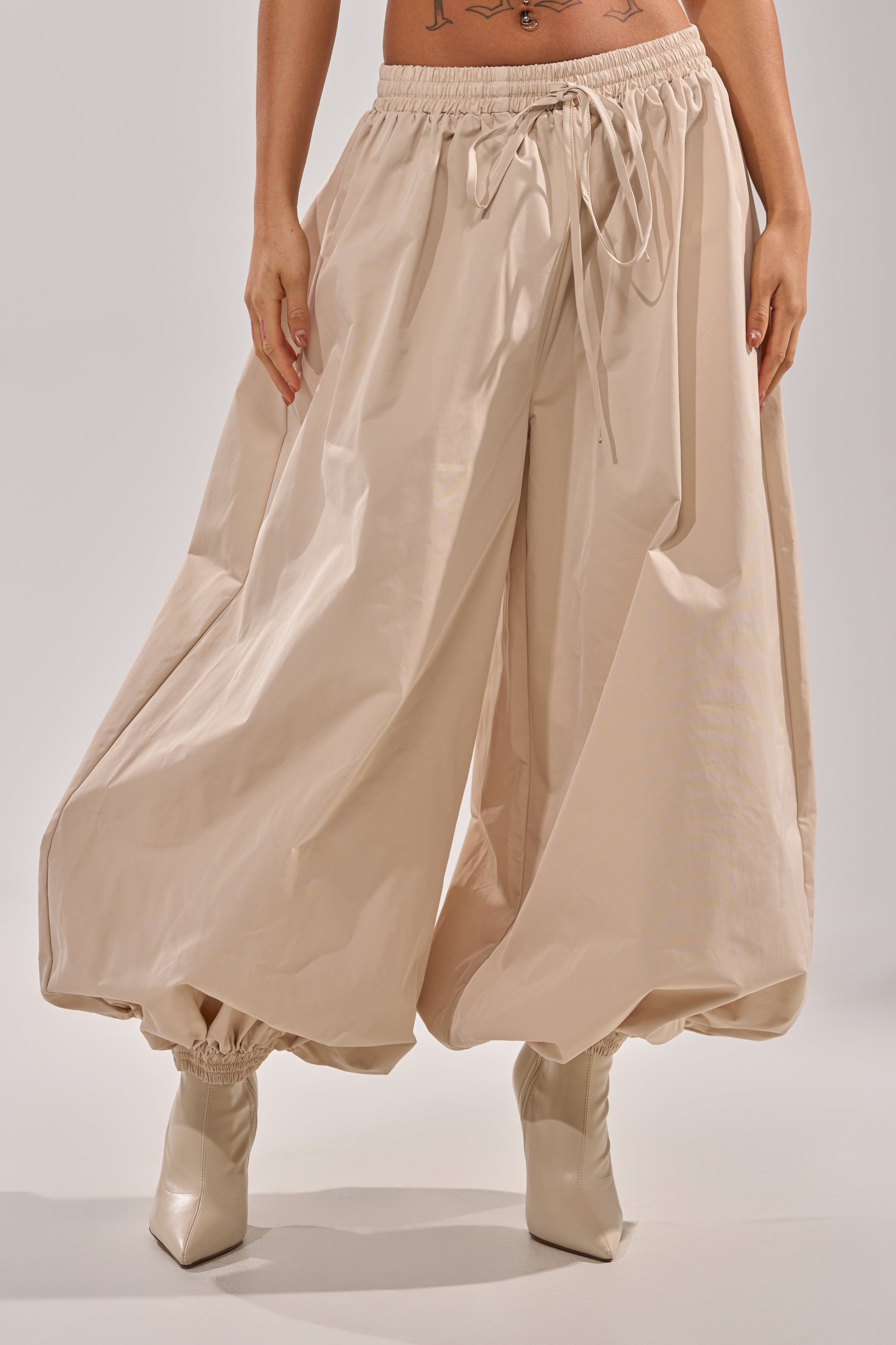 Wearing the GUESS WHO BALLOON PANT in beige with a relaxed high-rise fit, gathered ankles, and drawstring waist, paired with pointed-toe light boots. Only the lower half of this street style outfit is shown. 1