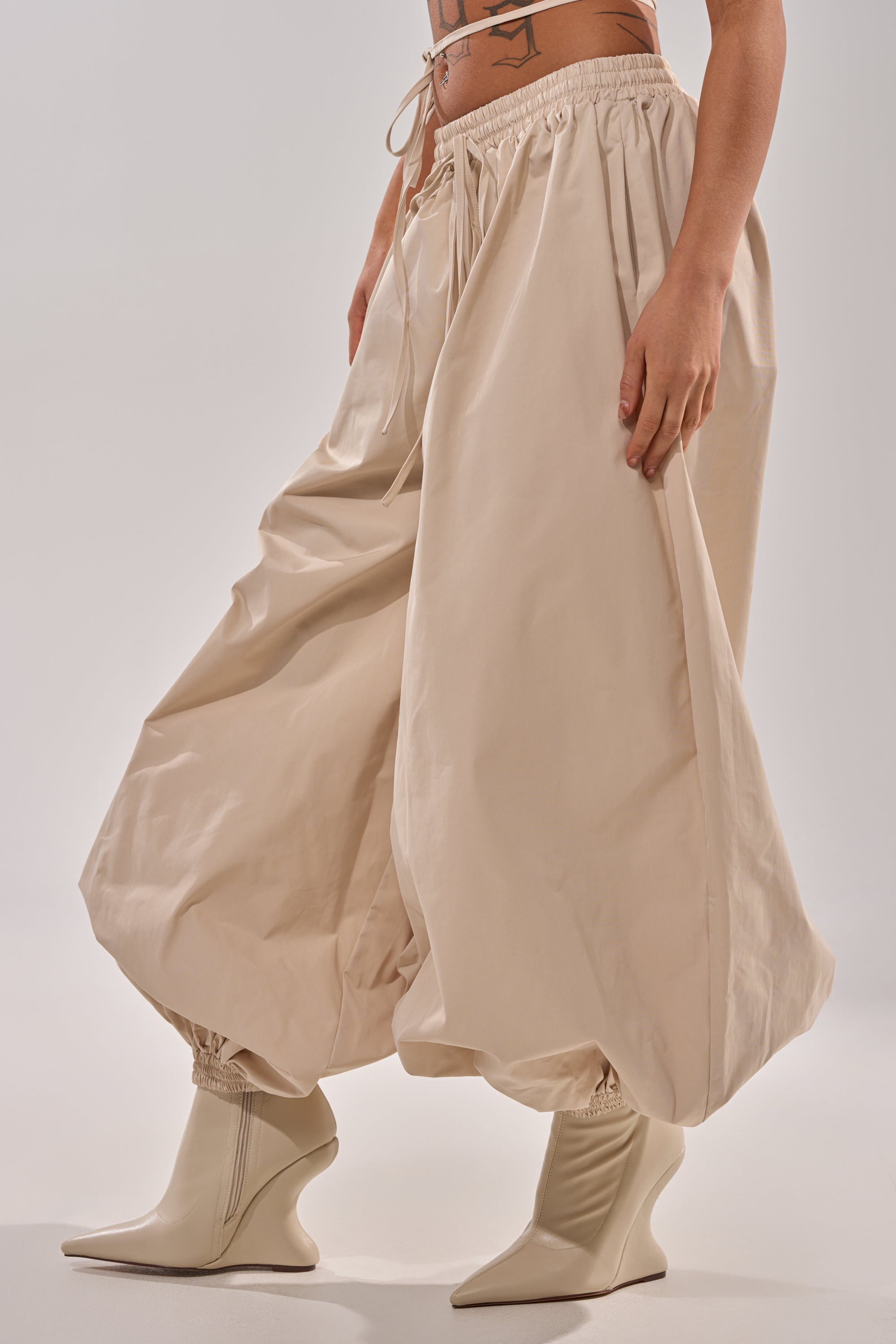 Someone shows off street style in the GUESS WHO BALLOON PANT—oversized, beige pants with an elastic waistband—paired with pointed, cream high-heeled boots against a neutral background. 3