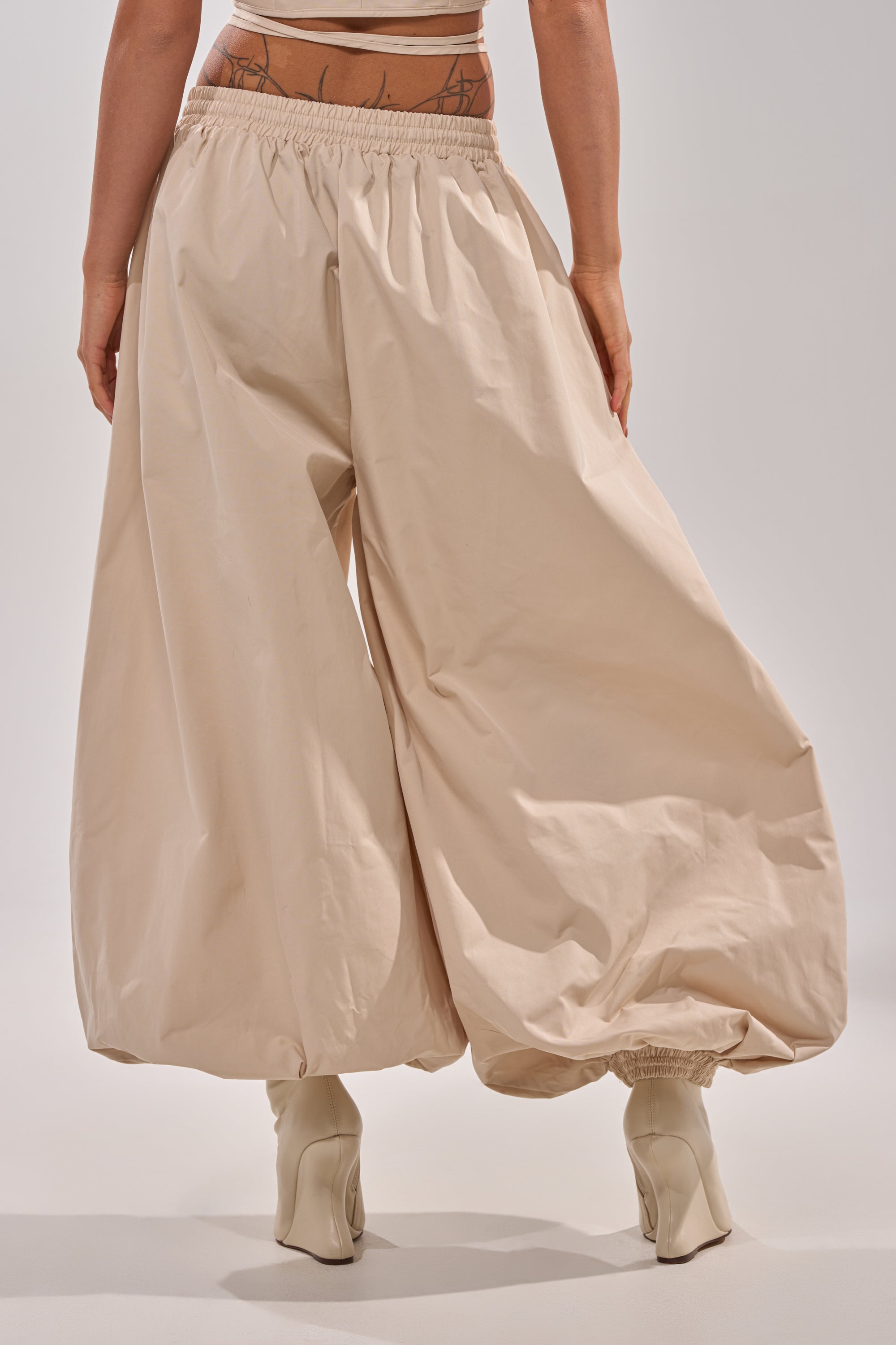 A person models the GUESS WHO BALLOON PANT—loose, beige with a relaxed high-rise fit and elastic cuffs—paired with white heeled boots, seen from the back against a plain light background. 4