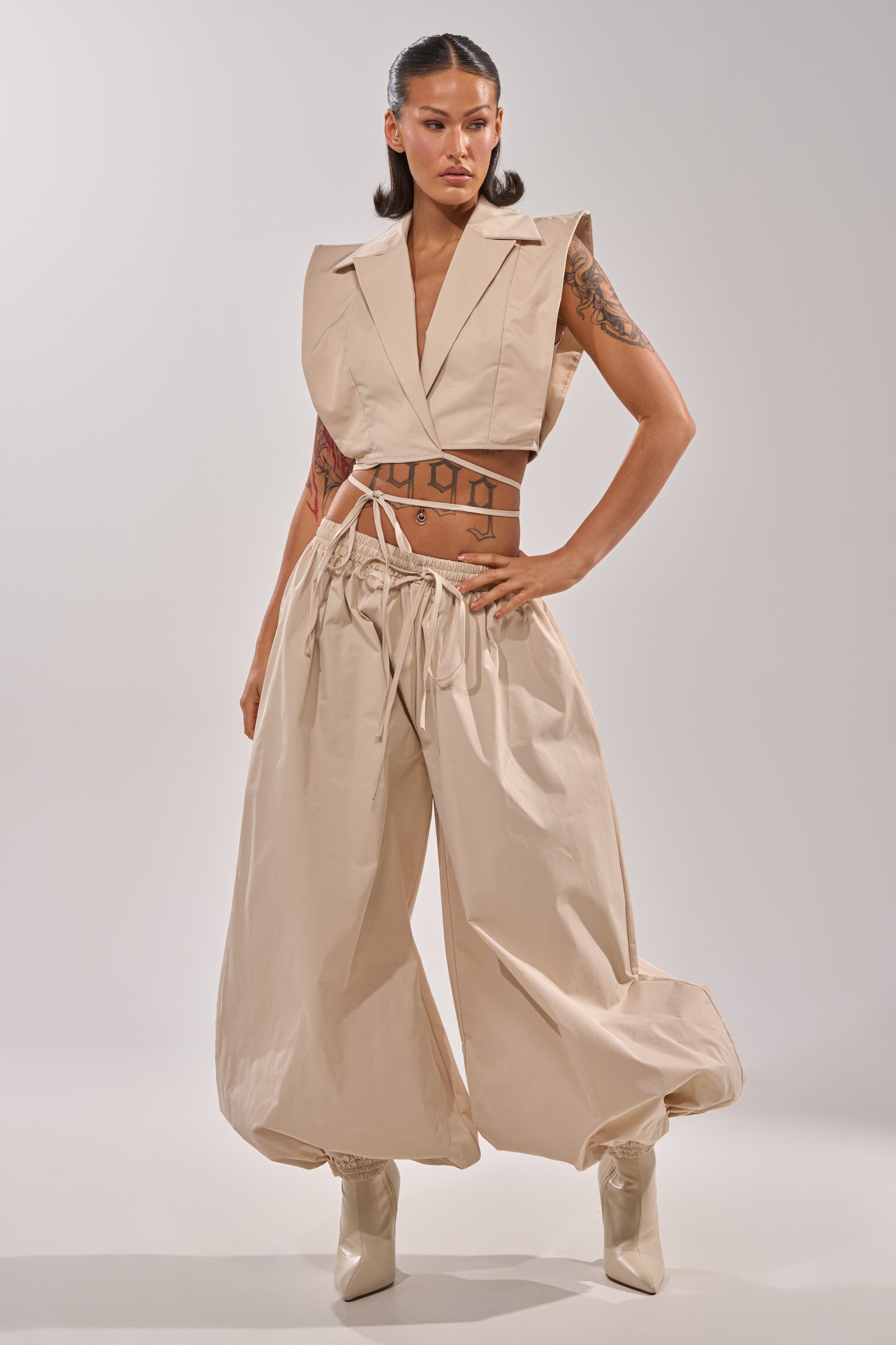 A woman models the GUESS WHO BALLOON PANT, featuring a relaxed high-rise fit. She pairs it with a beige sleeveless cropped vest, pointed-toe boots, and wears her sleek shoulder-length hair simply against a plain light background. 5