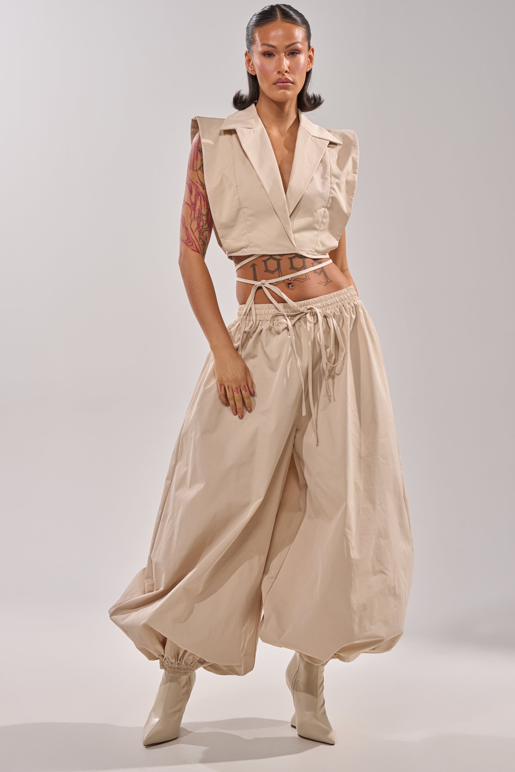 A woman with tattoos models the GUESS WHO BALLOON PANT—beige, high-rise, loose-fitting with a drawstring waist—styled with a cropped vest and pointed-toe boots for a trendy street style look. 6