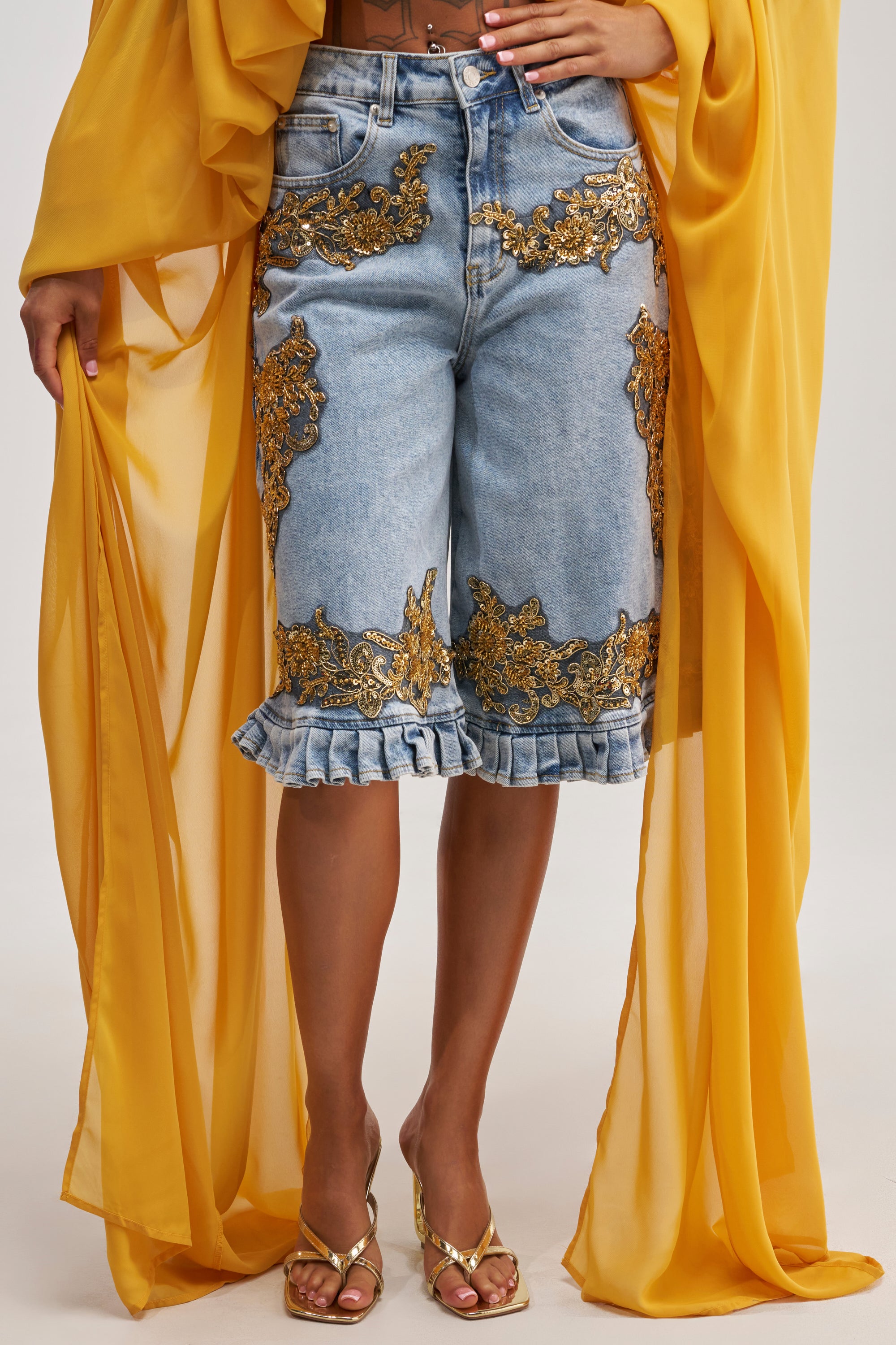 Wearing the BABY THAT’S HOW I LIKE IT BERMUDA SHORT—light-wash denim bermuda shorts featuring gold embroidery, embellishments, and ruffled hems—paired with gold heeled sandals and a flowing yellow sheer garment draped over the arms.