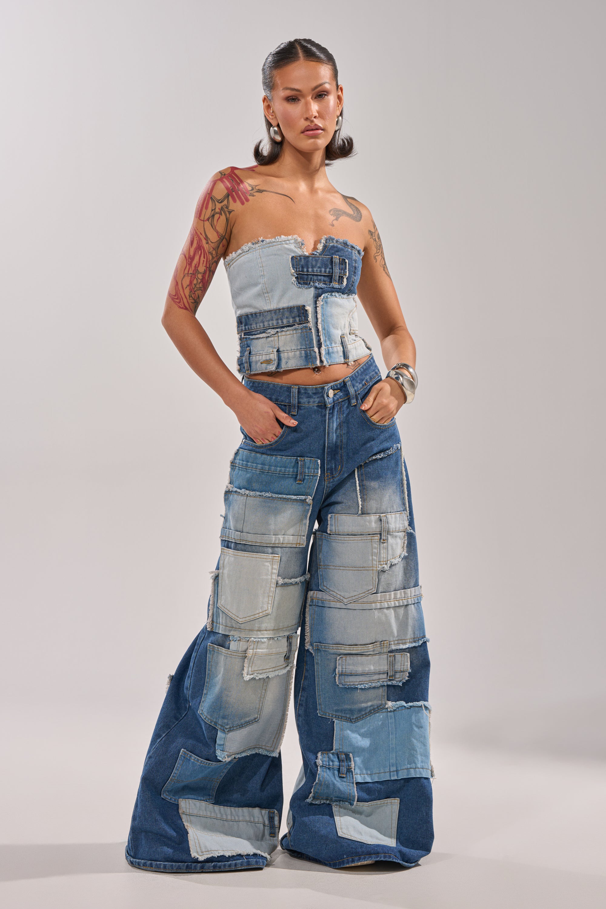 A woman with styled hair and tattoos stands confidently in PATCH ME UP RELAXED DENIM, wearing a strapless patchwork top and wide-leg pants, her hands by her waist against a plain background. 0