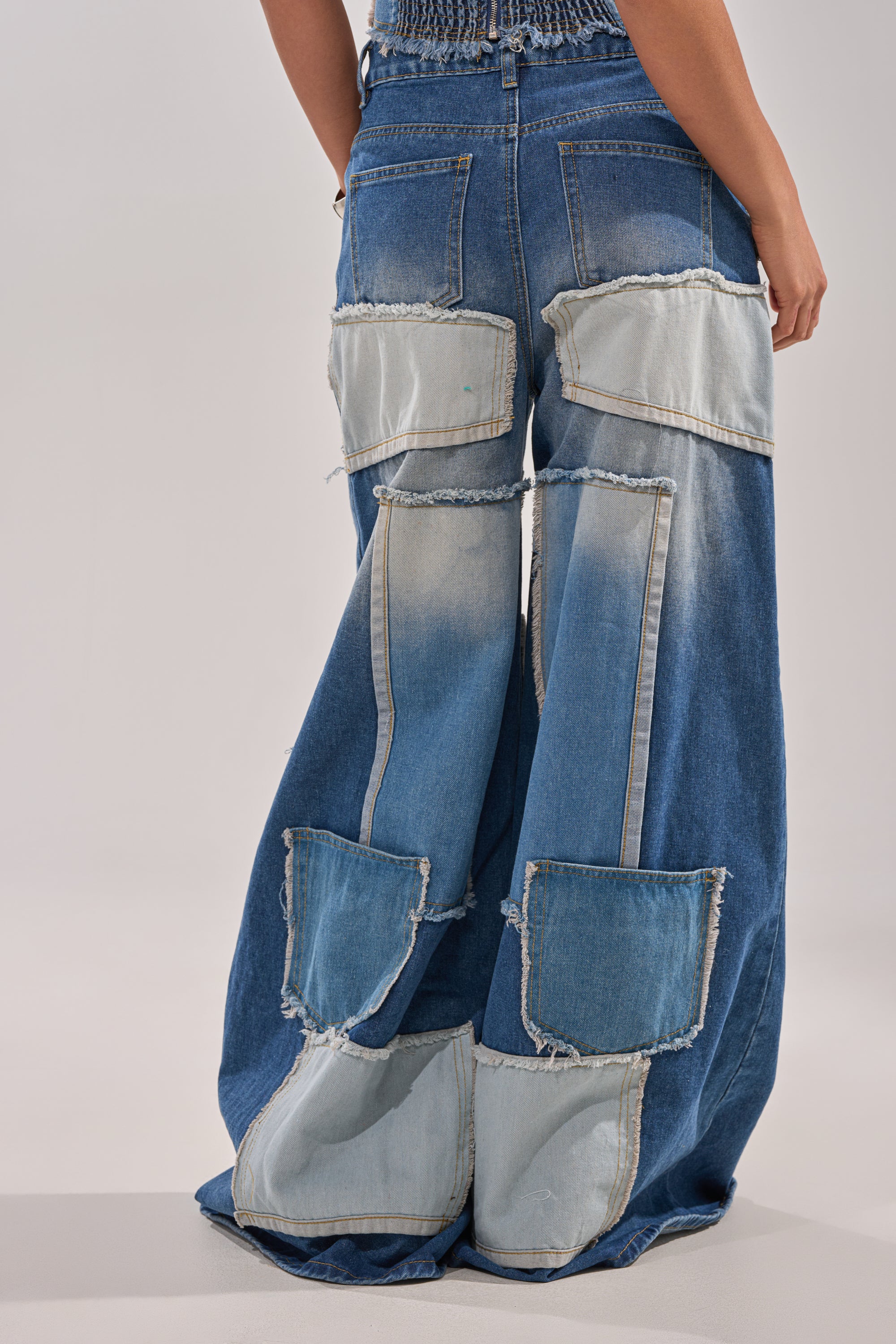 Someone wearing the PATCH ME UP RELAXED DENIM, featuring wide legs and large, frayed patchwork in varied denim shades on the back and legs, is shown from behind against a neutral background, channeling a bold streetwear style. 3