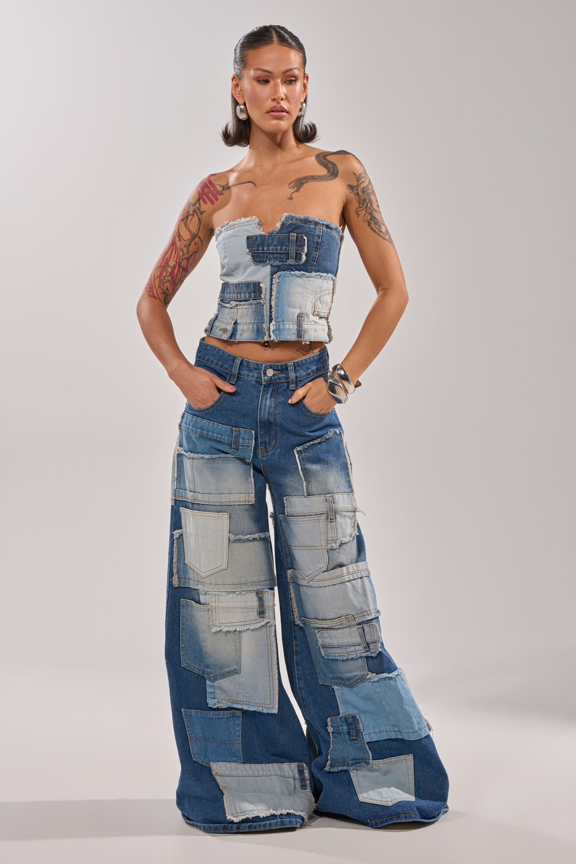 A woman with tattoos, hoop earrings, and long dark hair stands against a plain light background, hands in pockets, wearing the PATCH ME UP RELAXED DENIM—a strapless patchwork top and high-rise wide-leg jeans. 4