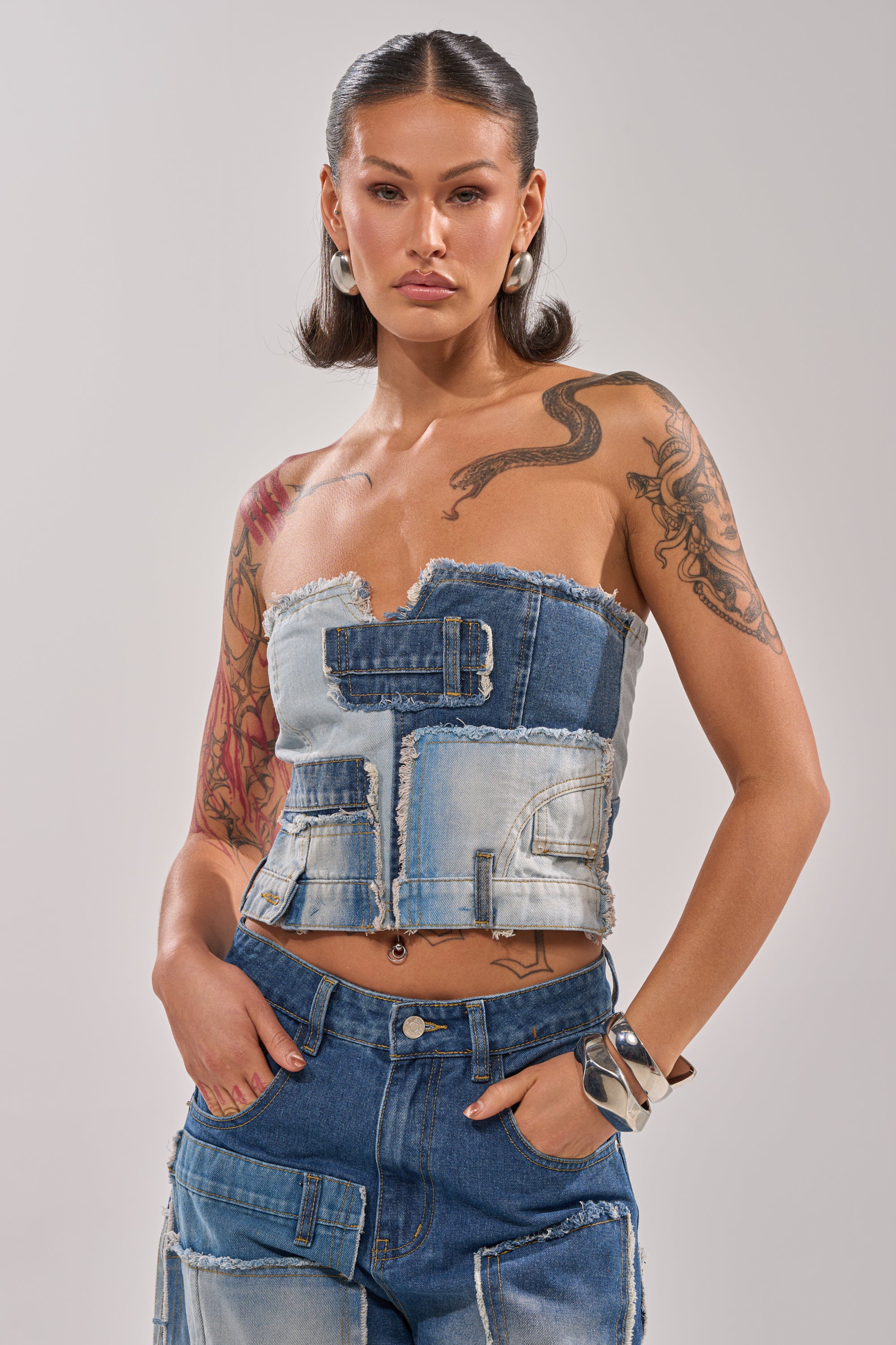 A tattooed woman with short hair, hoop earrings, and a metal bracelet poses confidently in the PATCH ME UP DENIM CORSET and matching patchwork jeans against a plain background. 0