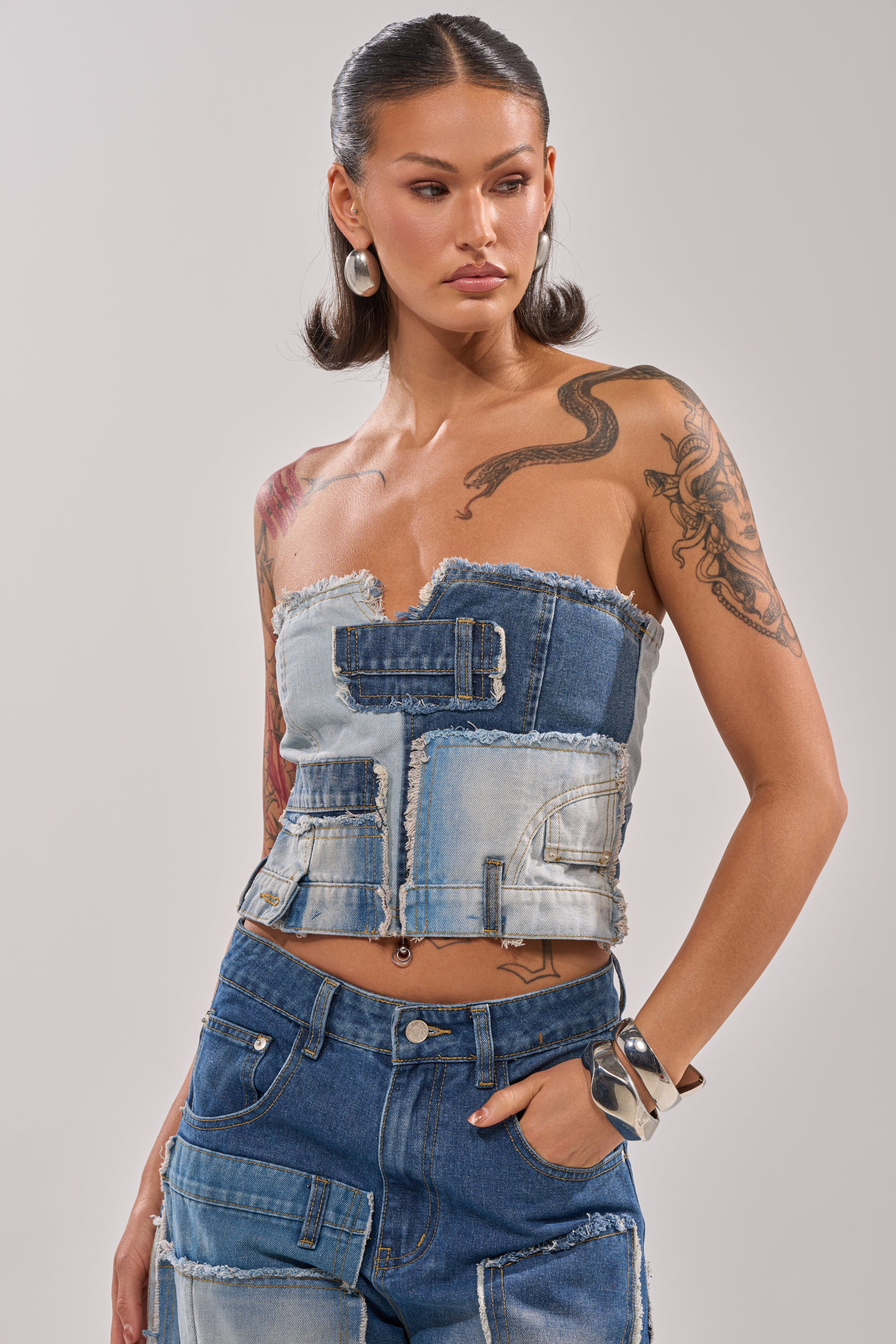 A woman with dark wavy hair wears the PATCH ME UP DENIM CORSET and matching jeans, showcasing Y2K style with visible pockets and seams. She displays arm tattoos, silver jewelry, and poses against a plain background. 1