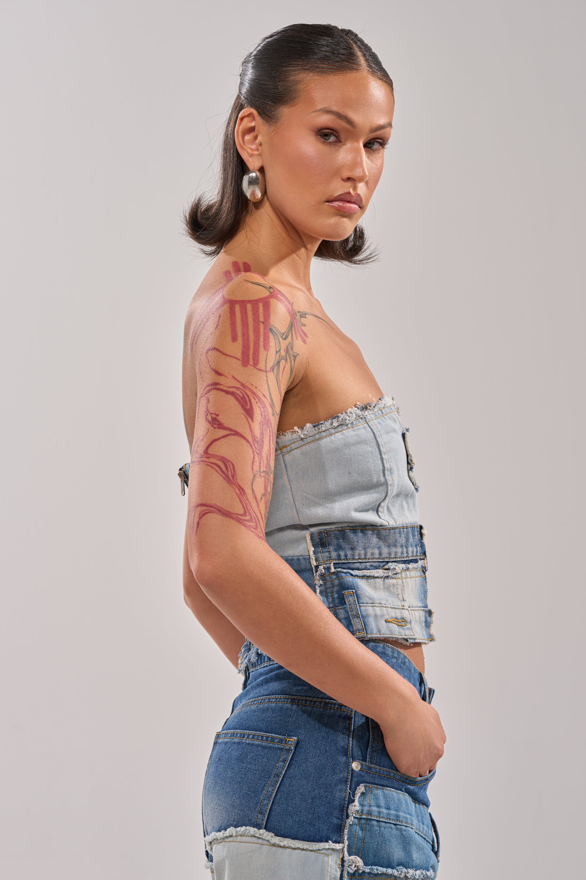 A woman with dark, sleek hair and a bold upper arm tattoo wears the PATCH ME UP DENIM CORSET. Paired with jeans and silver earrings, she stands confidently against a plain backdrop, channeling Y2K-inspired patchwork style. 2