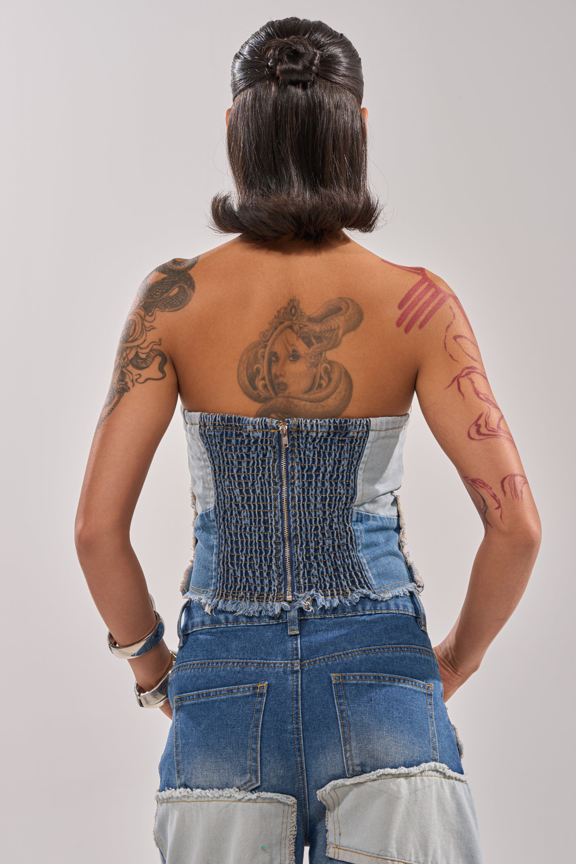 A person with short, styled dark hair and visible tattoos wears the PATCH ME UP DENIM CORSET and high-waisted jeans, standing against a plain light gray background. 3
