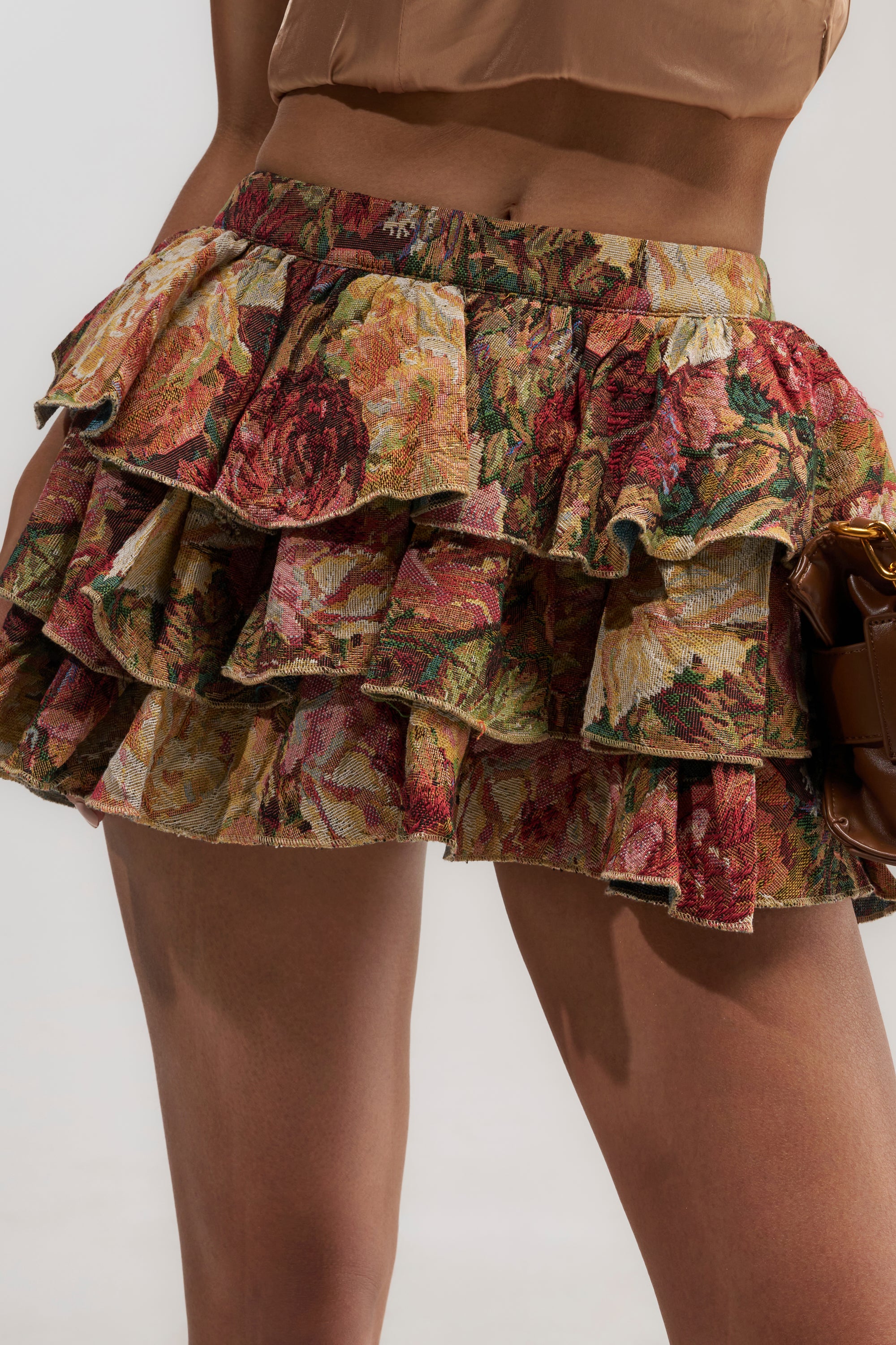 A person wearing the WILD FLORAL TAPESTRY SKIRT styled with a brown cropped top and holding a small brown bag, set against a plain light-colored background.