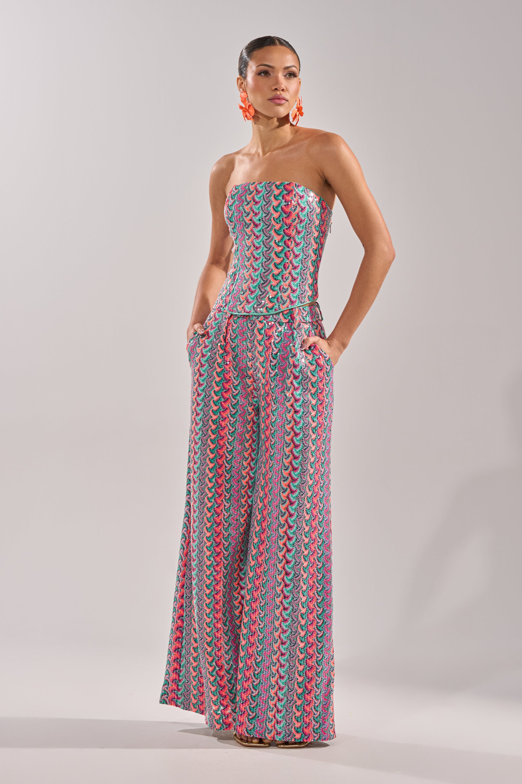 A woman wears the COASTAL BABE TROUSER, a colorful, strapless jumpsuit with wide-leg pants and a blue, pink, and yellow wavy pattern—ideal for vacations. She completes the look with bright orange earrings and slicked-back hair. 3