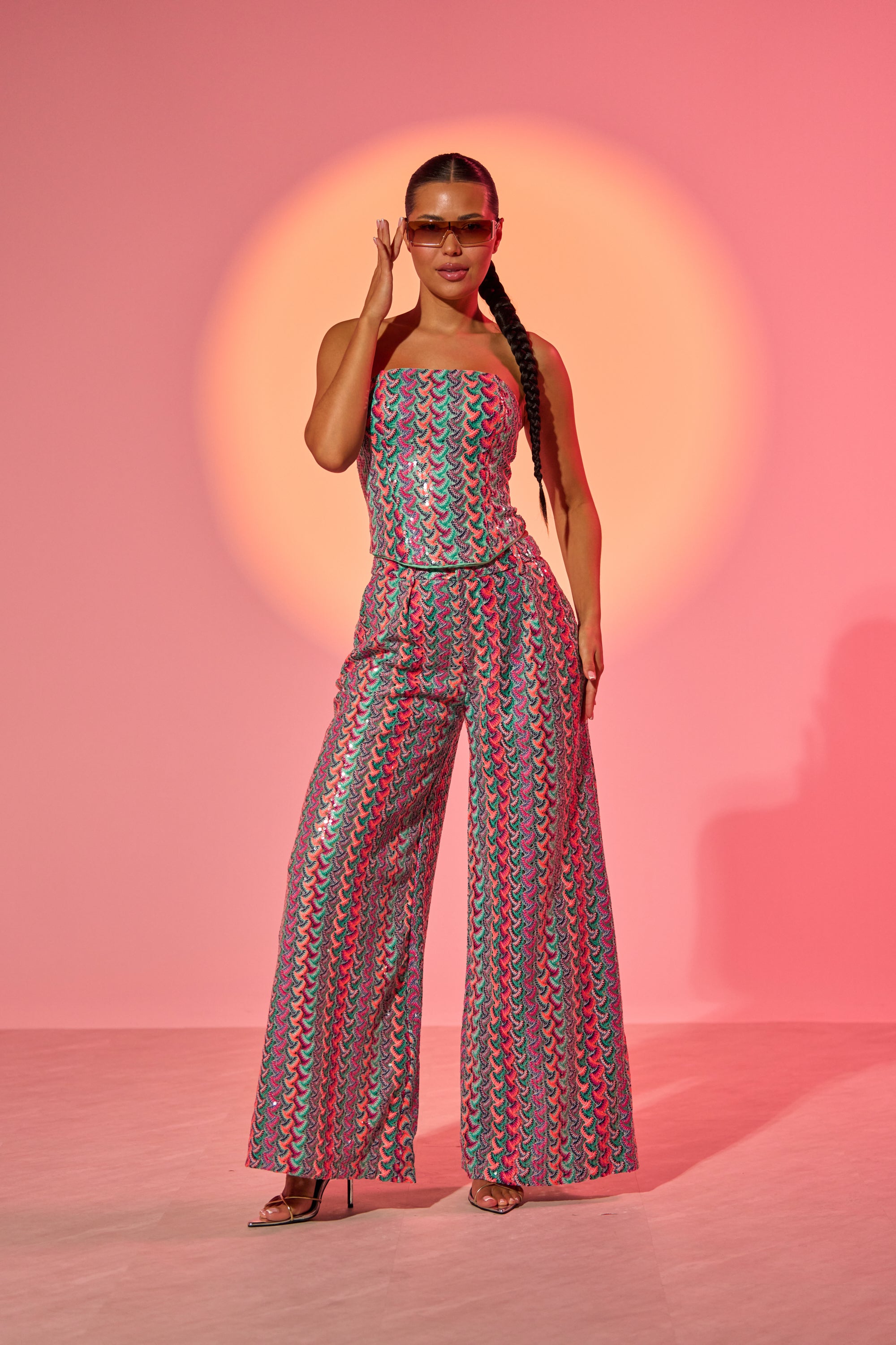A woman exudes confidence in the COASTAL BABE TROUSER—high-waisted sequin pants in a vibrant, multicolored design—styled with sunglasses and heels against a pink backdrop with orange light, making it perfect for standout vacation looks. 0