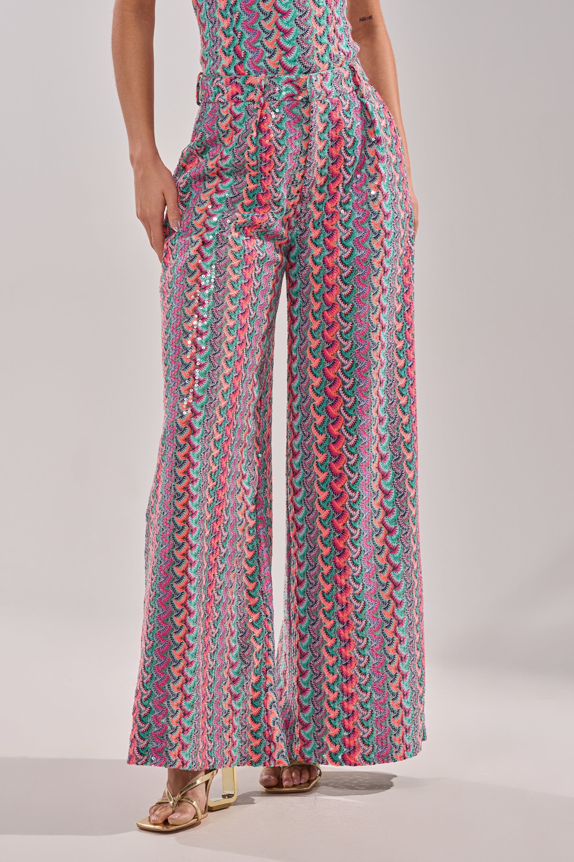 A person wears the COASTAL BABE TROUSER—vibrant, wide-leg pants with a colorful pink, teal, and white wavy geometric pattern—paired with gold open-toed heels against a plain background, perfect for vacation style. 4