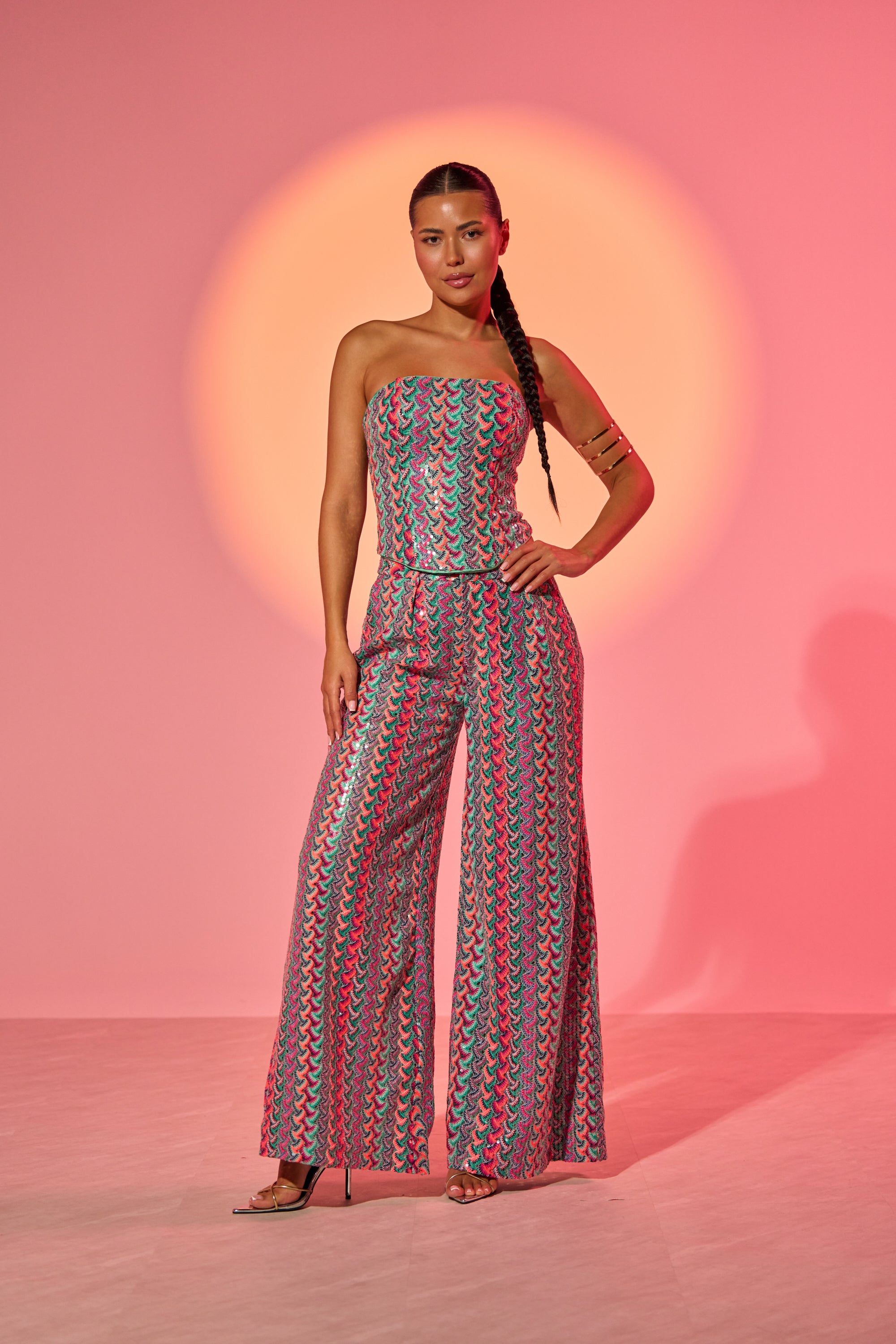 A woman poses confidently against a pink backdrop with a circular light pattern, wearing the COASTAL BABE TROUSER—a vibrant, wide-leg sequin pant that's perfect for standout vacation style. Her hair is styled in a sleek, long braid. 1