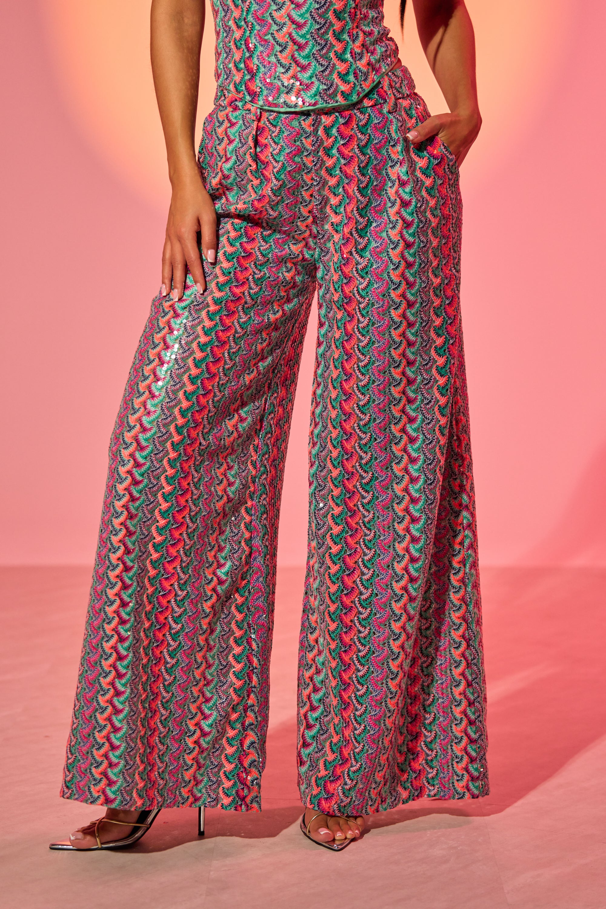 Against a pink background, a person poses with one hand in their pocket, wearing the COASTAL BABE TROUSER—colorful, patterned wide-leg sequin pants ideal for statement vacation style. Only their body from shoulders down is visible. 2