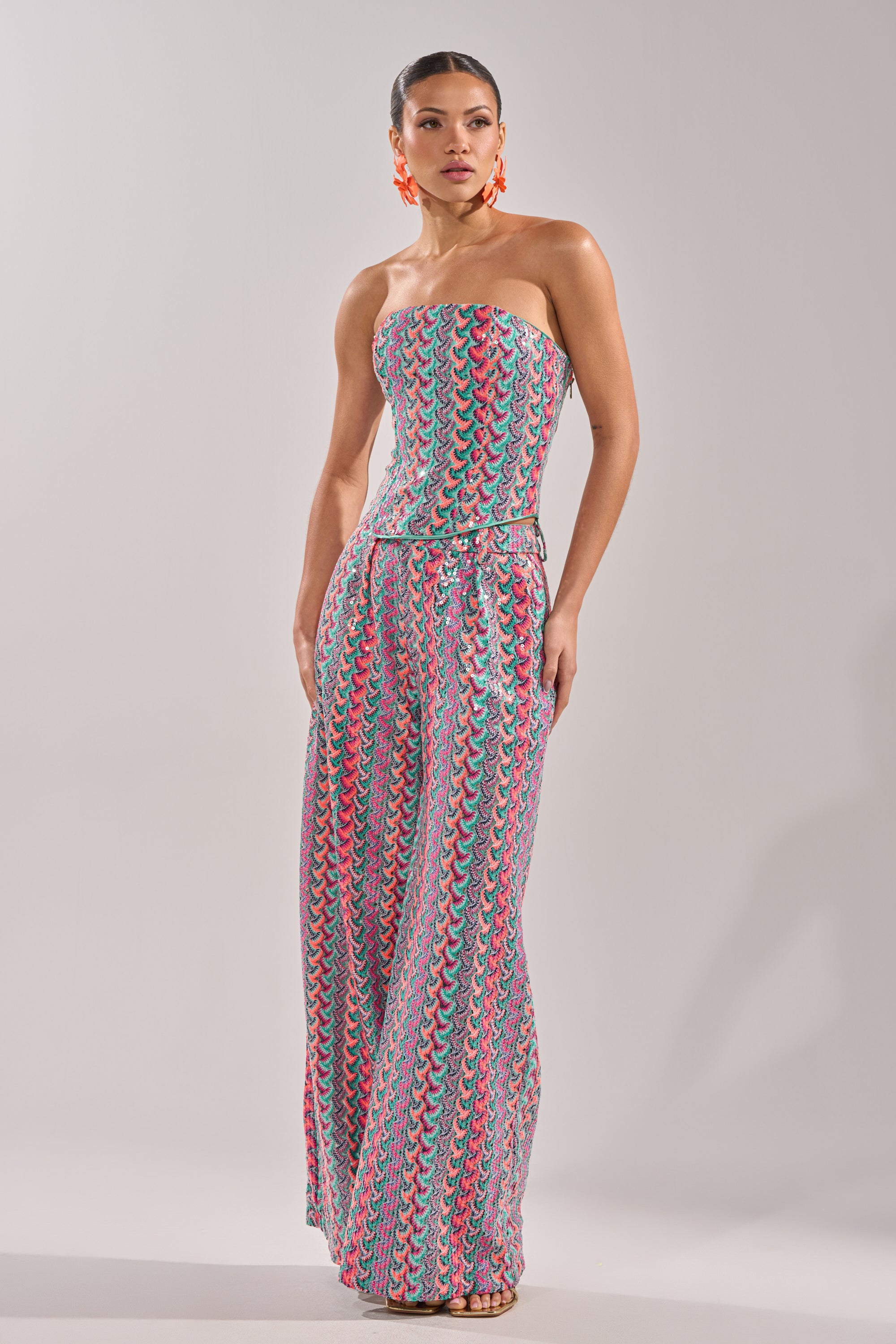 A woman models the COASTAL BABE TROUSER, a strapless, colorful patterned wide-leg jumpsuit ideal for vacations. She pairs it with bold orange earrings and wears her hair sleeked back against a plain light-colored background. 8