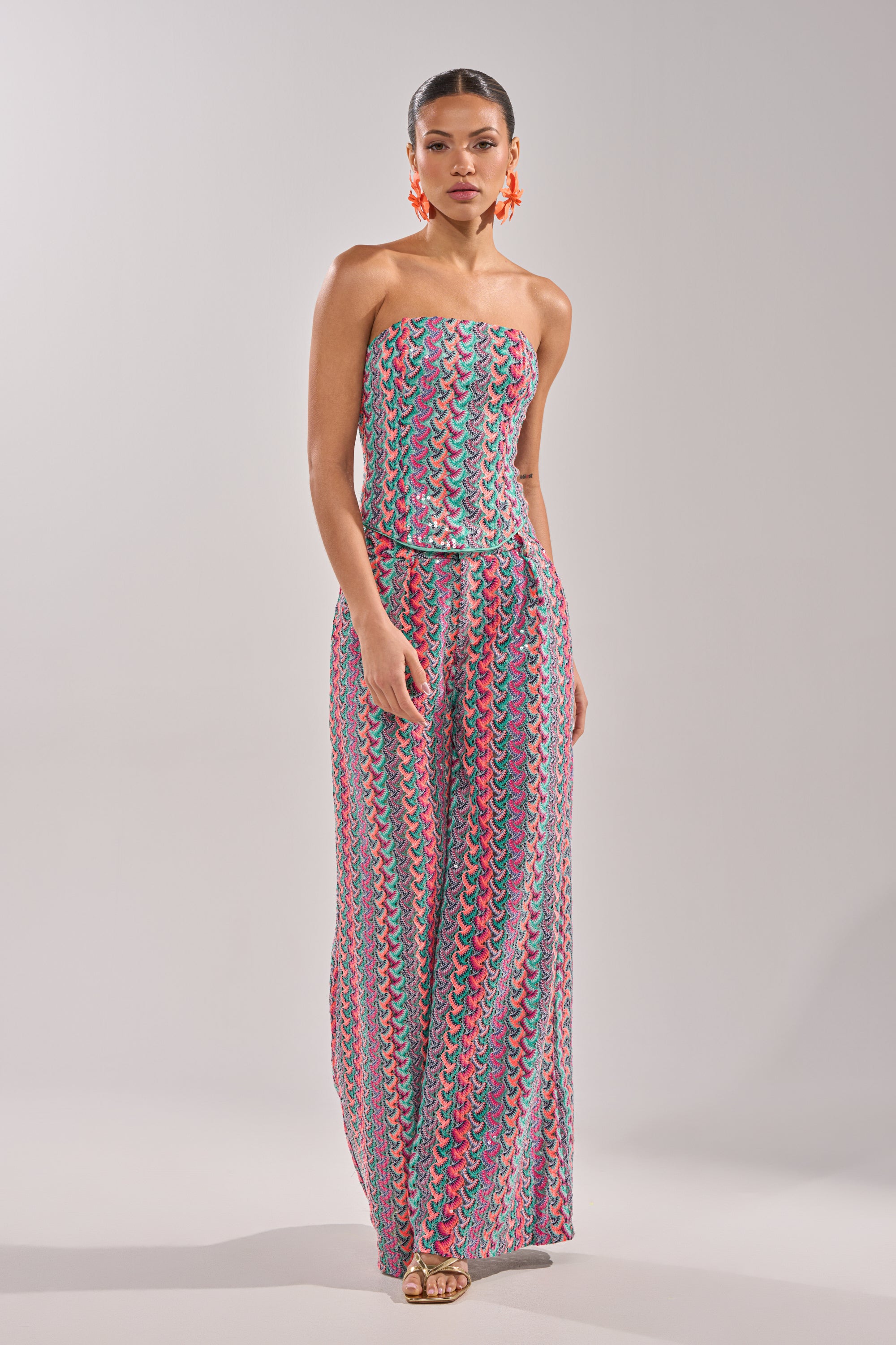 A woman poses against a plain light background wearing the COASTAL BABE TROUSER, a strapless jumpsuit with a colorful wavy pattern in pink, teal, and white. She accessorizes with bold orange earrings and gold open-toe heels—perfect for vacation. 9