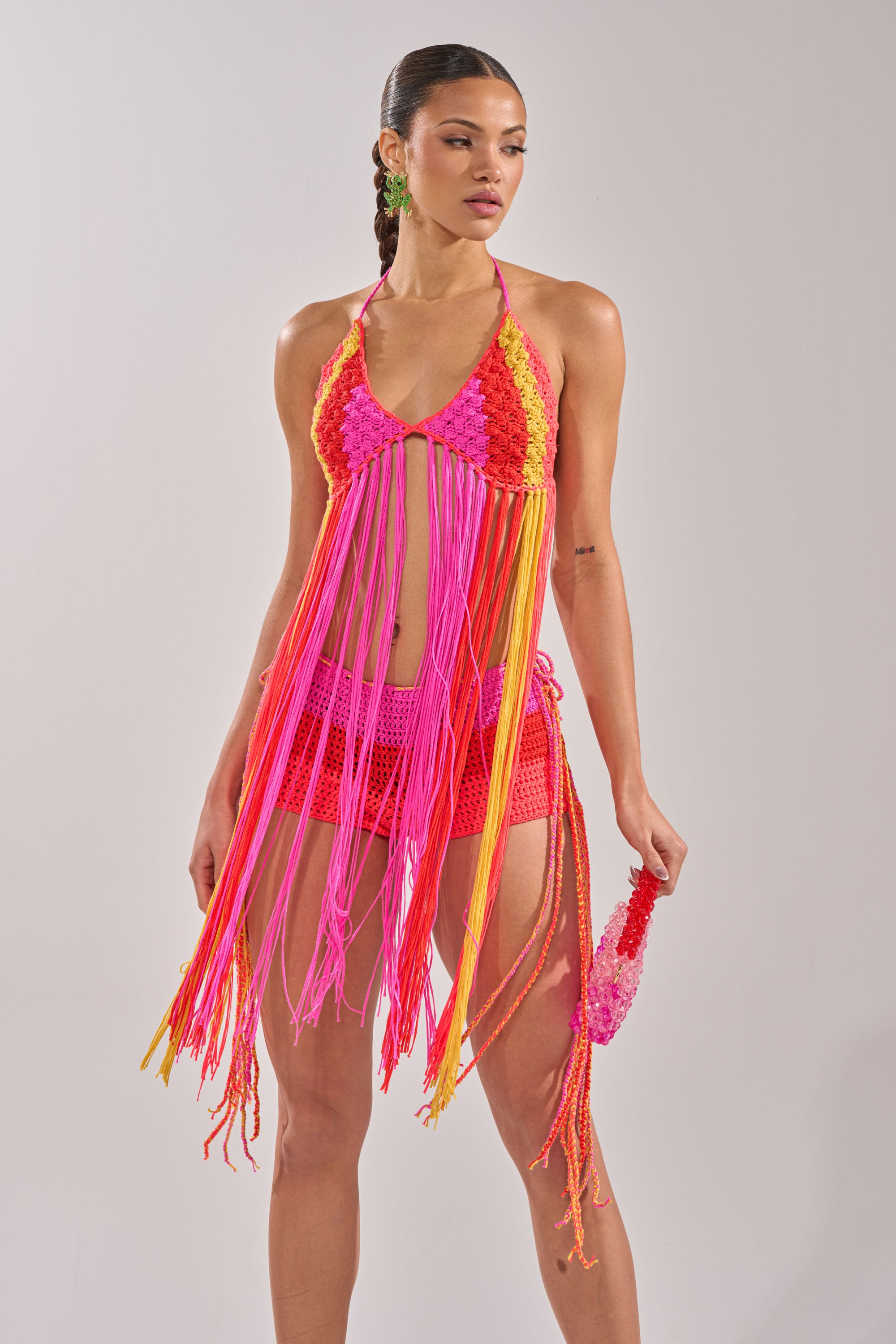 A woman poses in the BEACHIN CROCHET BRALETTE, a pink and orange crochet top with fringe, paired with matching shorts and a small crochet accessory, against a plain light background. 2