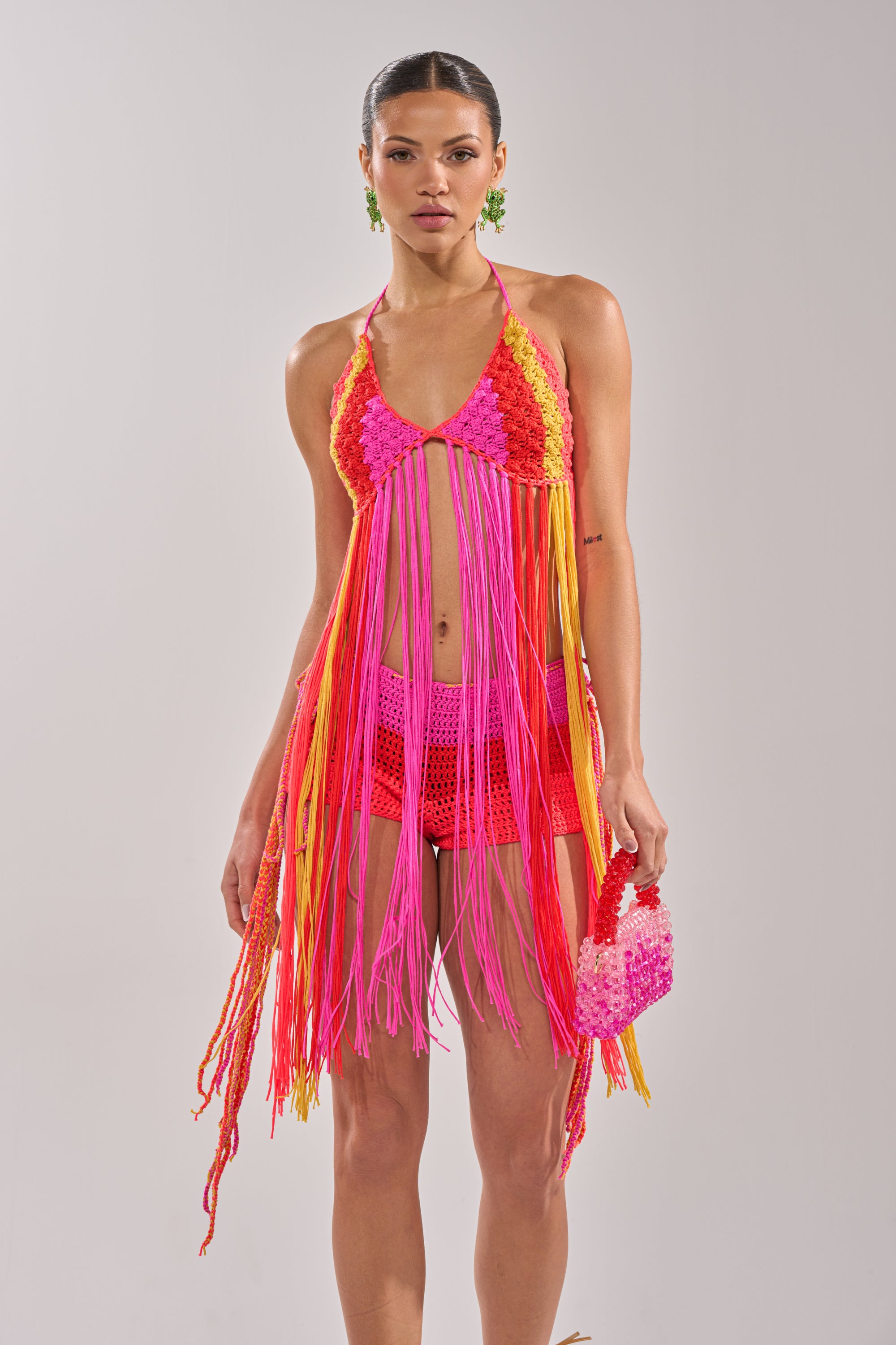 A woman wears the BEACHIN CROCHET BRALETTE, featuring colorful crochet with long pink, orange, and yellow fringe, matching shorts, green earrings, and a small pink and white handbag against a plain light background. 3
