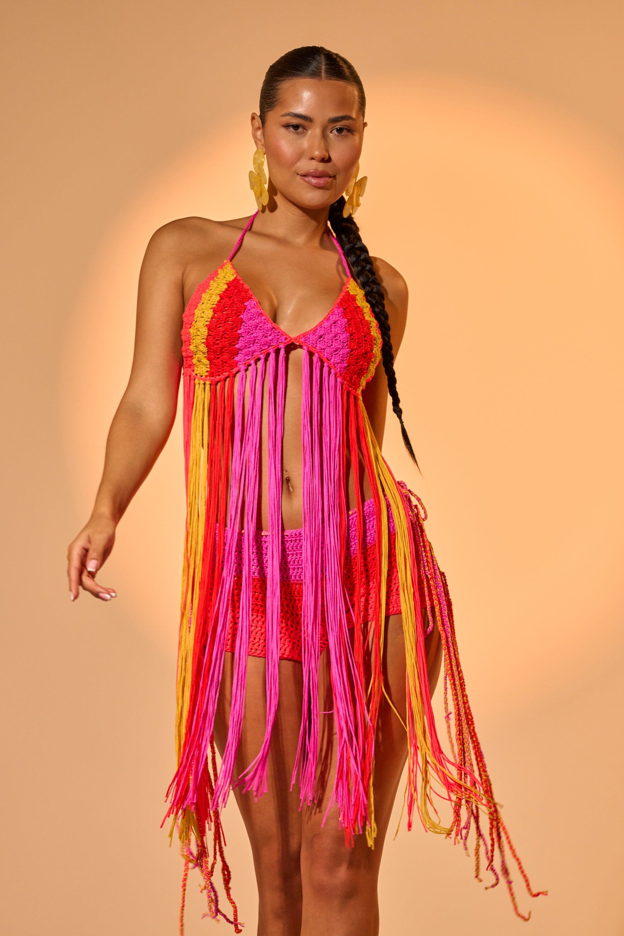 A woman poses against a beige background, wearing the BEACHIN CROCHET BRALETTE and a matching pink, orange, and yellow skirt with long fringe. She has long braided hair and statement earrings. 1