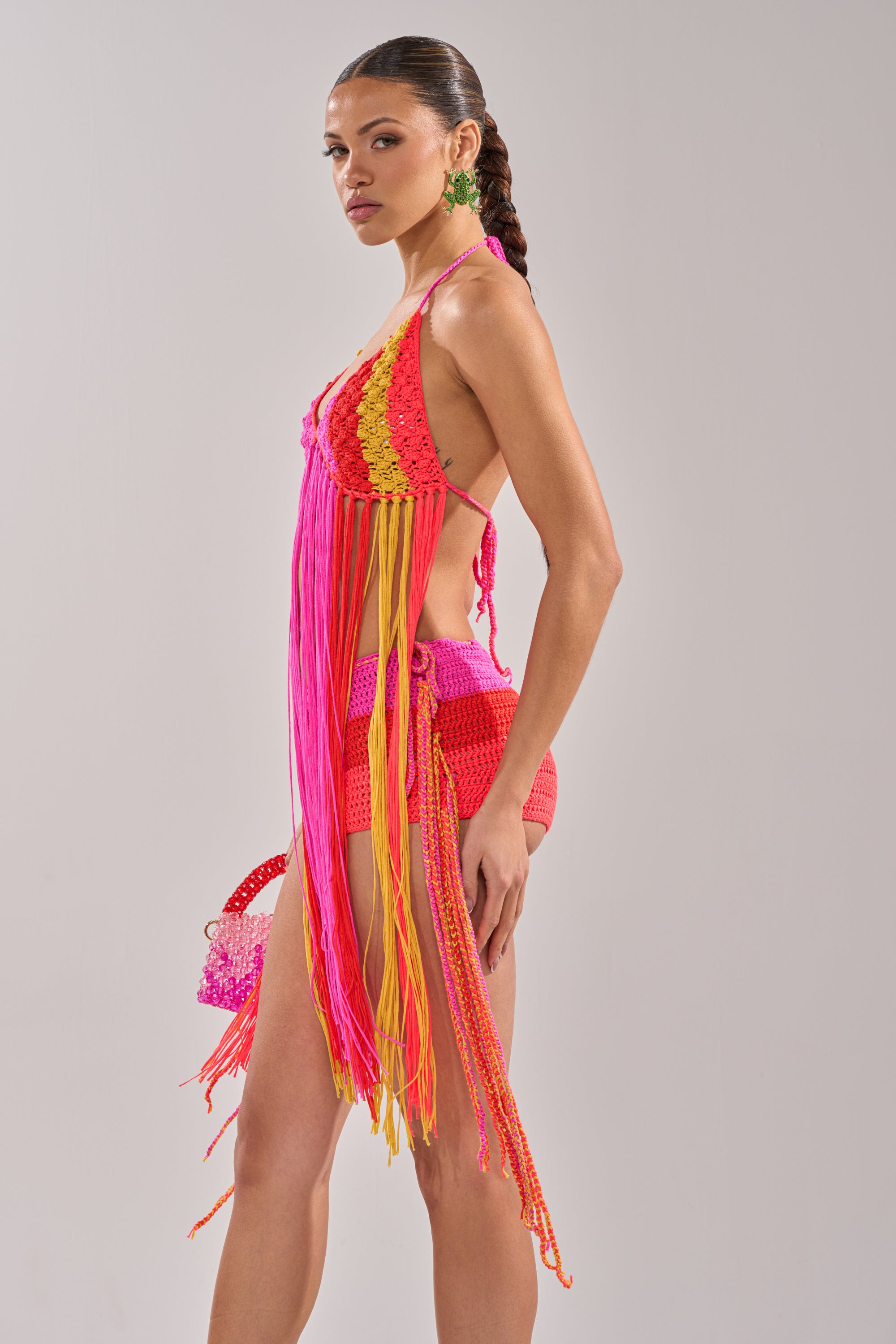 A woman poses in the BEACHIN CROCHET BRALETTE with a matching colorful mini skirt, fringe details, dangling earrings, and a small pink beaded handbag against a plain light background. 5
