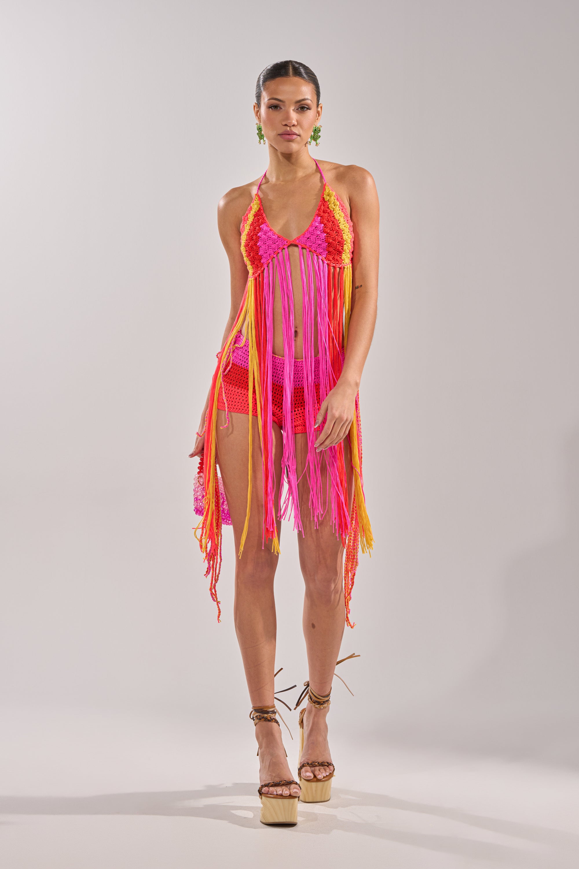 A woman wears the BEACHIN CROCHET BRALETTE in vibrant pink, orange, and yellow hues with fringe detail, paired with platform sandals and statement earrings. Her hair is tied back as she poses against a plain background. 8
