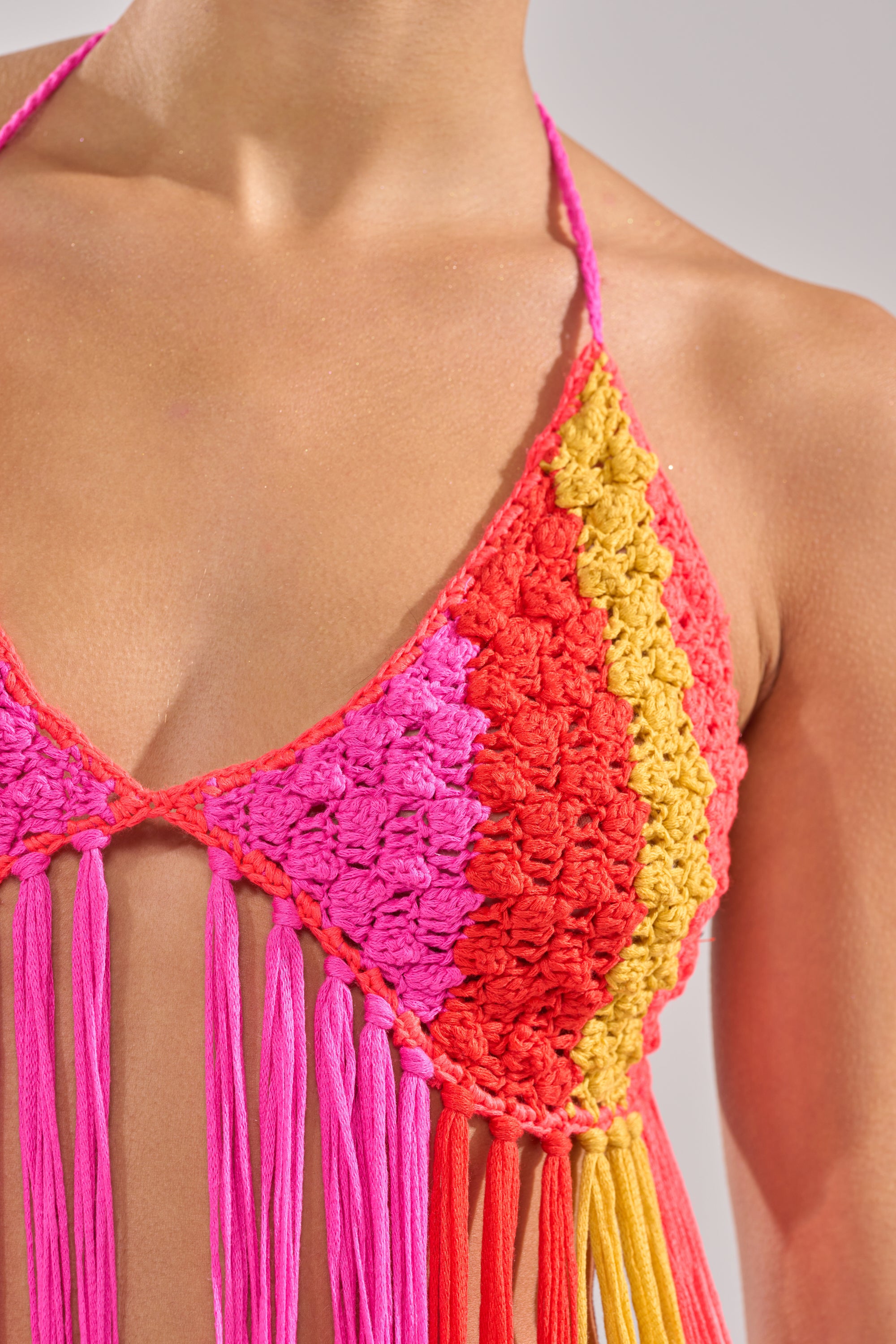 A close-up of a person wearing the BEACHIN CROCHET BRALETTE, featuring vibrant pink, red, and yellow sections with long fringe. The shot highlights the unique crochet style across the upper chest, shoulder, and neck. 9