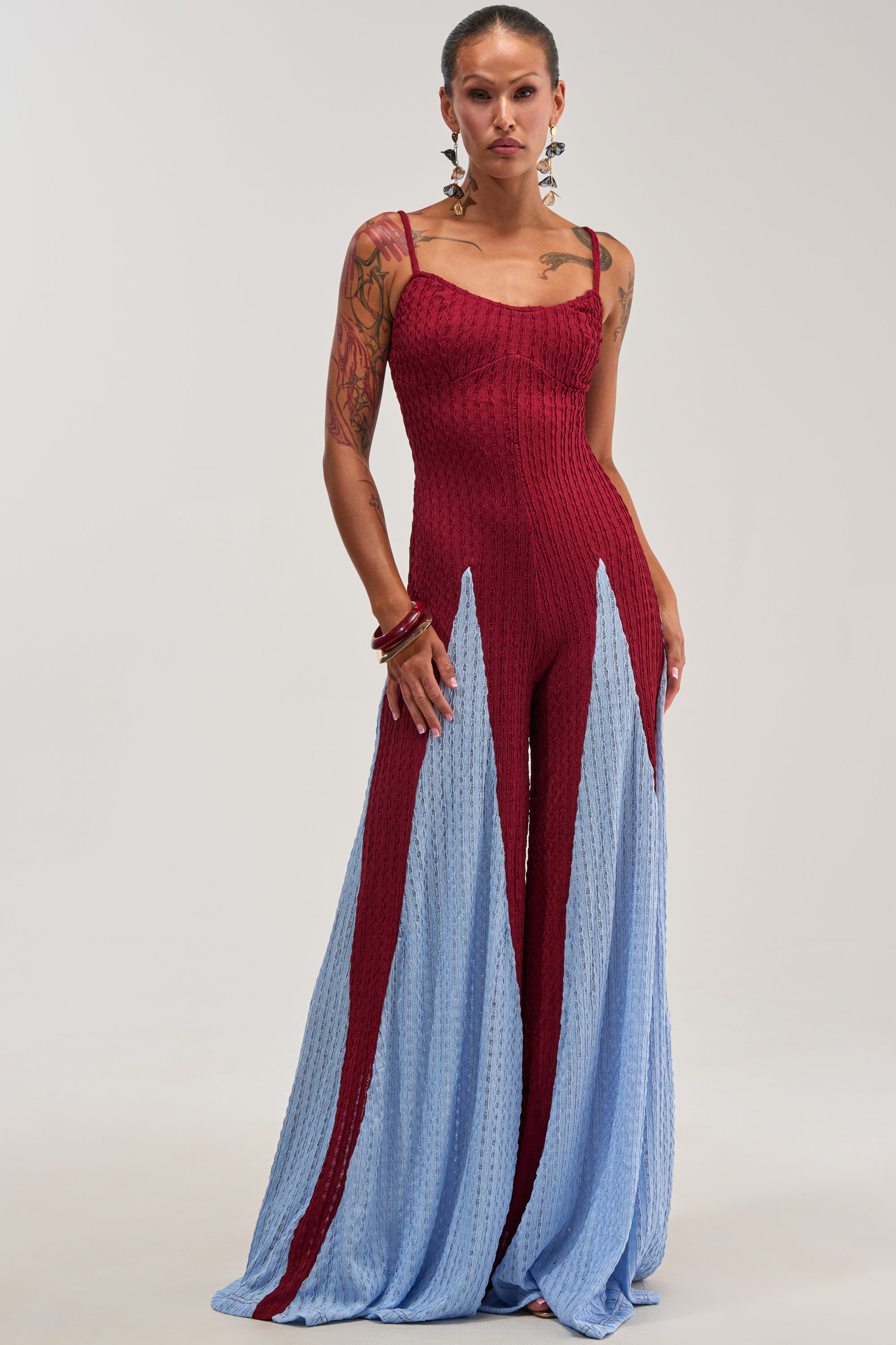 A person wearing the ALL DAY EVERYDAY COLOR BLOCK WOVEN JUMPSUIT IN BURGUNDY, a sleeveless jumpsuit featuring maroon and light blue color blocking with wide legs, stands against a plain light background. They have tattoos, earrings, and bracelets.