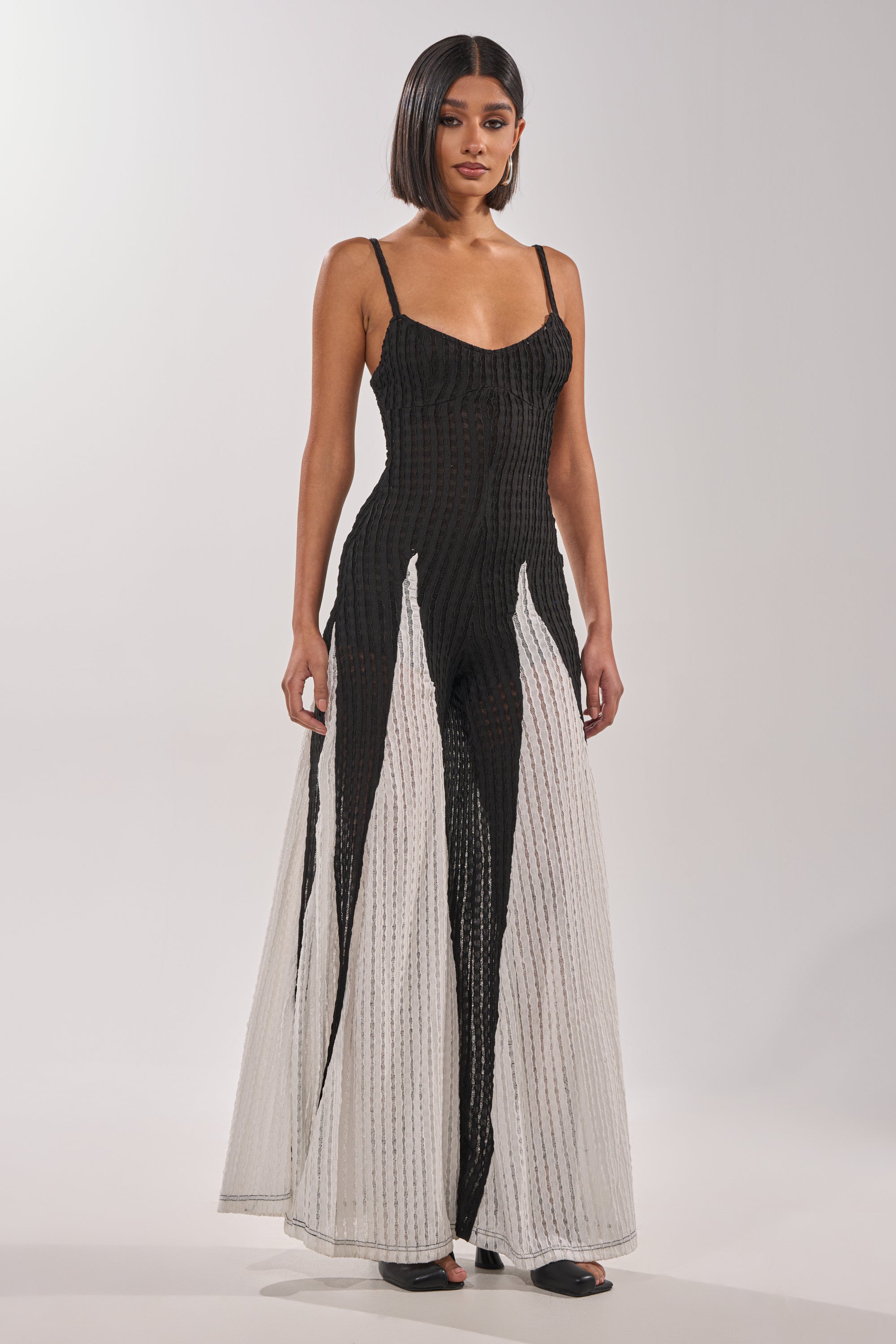 A woman with straight, shoulder-length hair models the ALL DAY EVERYDAY COLOR BLOCK WOVEN JUMPSUIT, which features a fitted black bodice and flared white panels from the knees down. She stands against a plain light background. 1