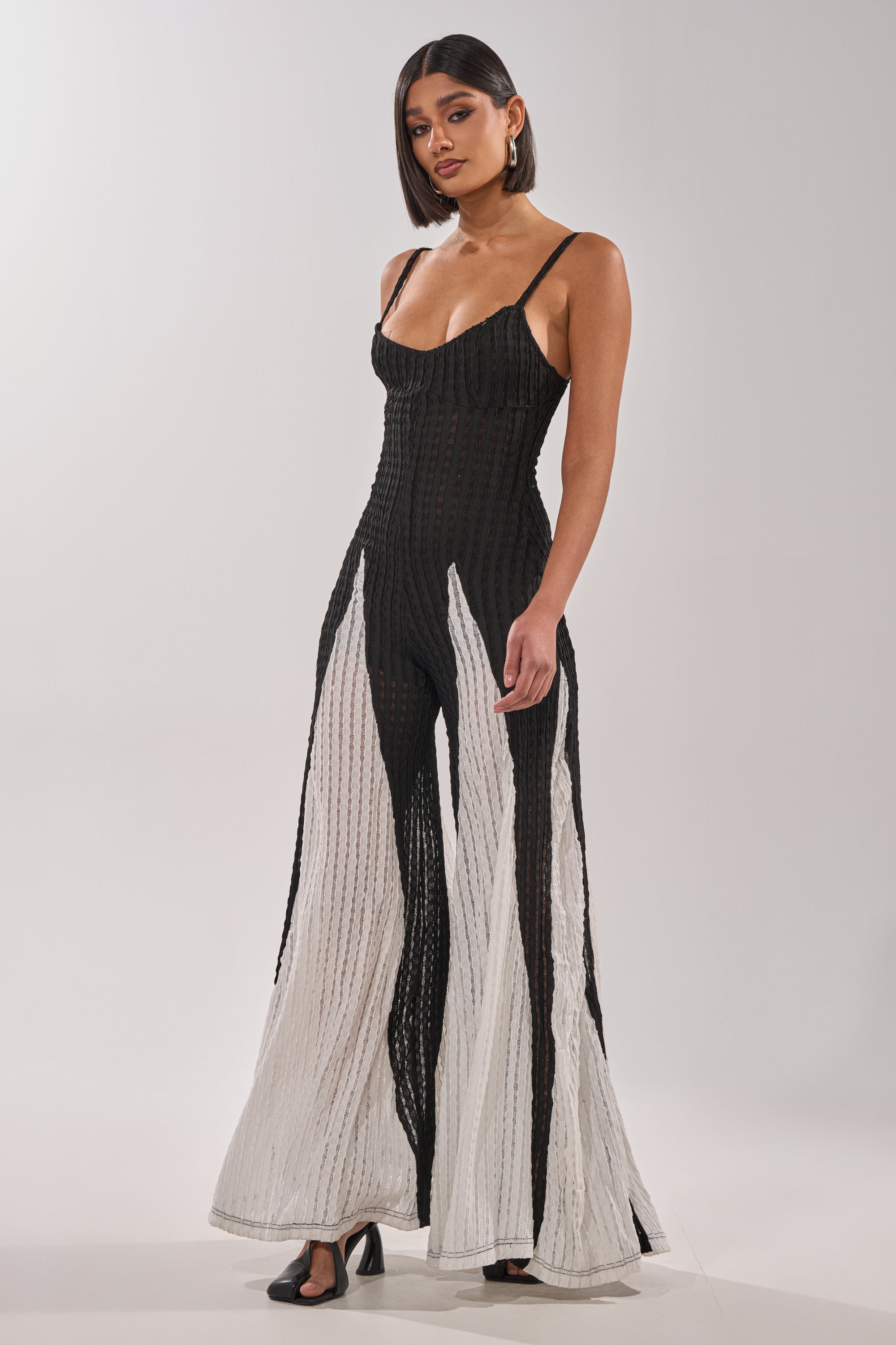 A woman with straight dark hair, hoop earrings, and black heels models the ALL DAY EVERYDAY COLOR BLOCK WOVEN JUMPSUIT—a sleeveless black and white wide-leg style—against a plain light background. 0