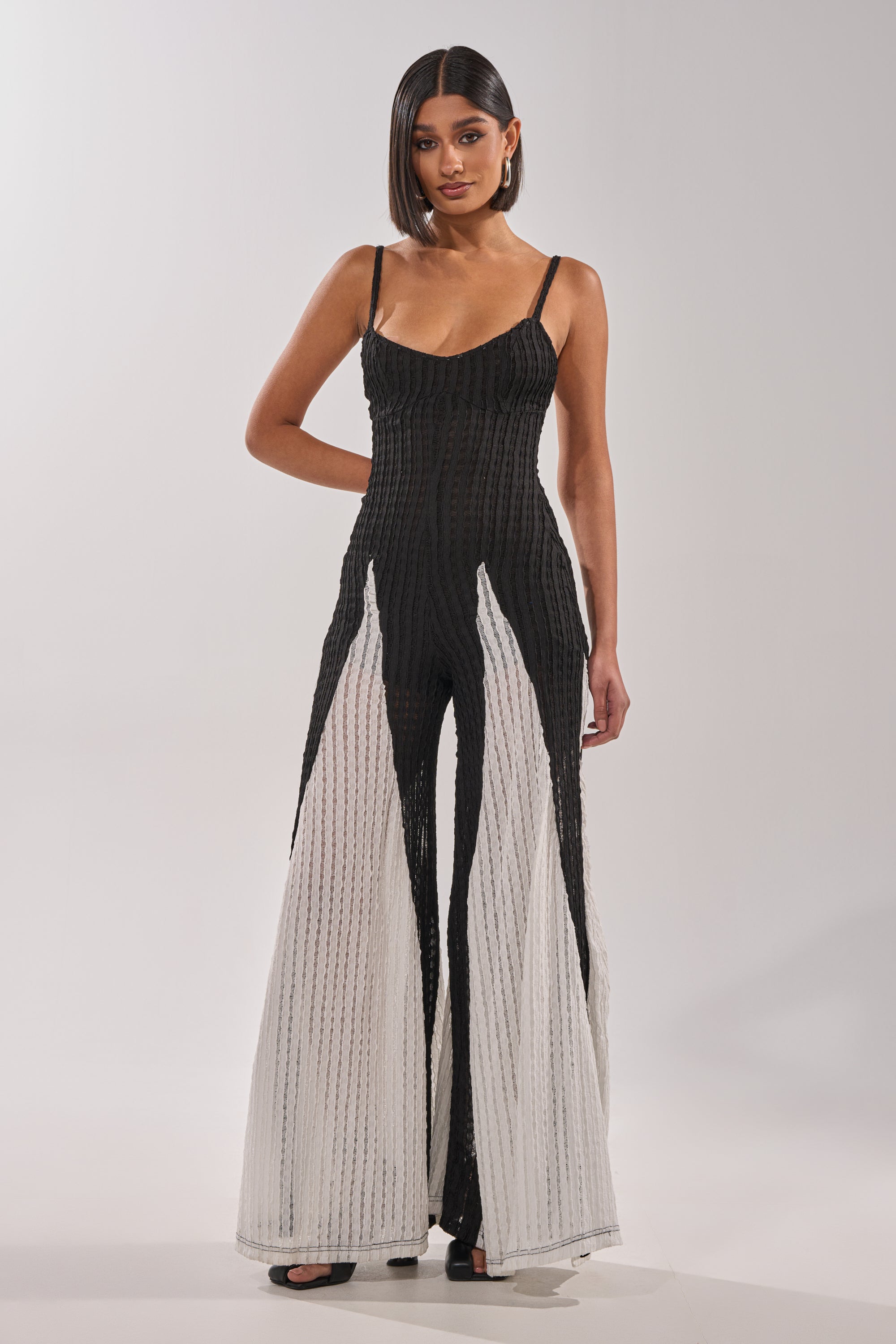 A woman poses confidently with one hand on her hip, wearing the ALL DAY EVERYDAY COLOR BLOCK WOVEN JUMPSUIT—a sleek black and white piece with wide-leg flared panels—against a plain light background. Her straight hair falls to her shoulders. 2