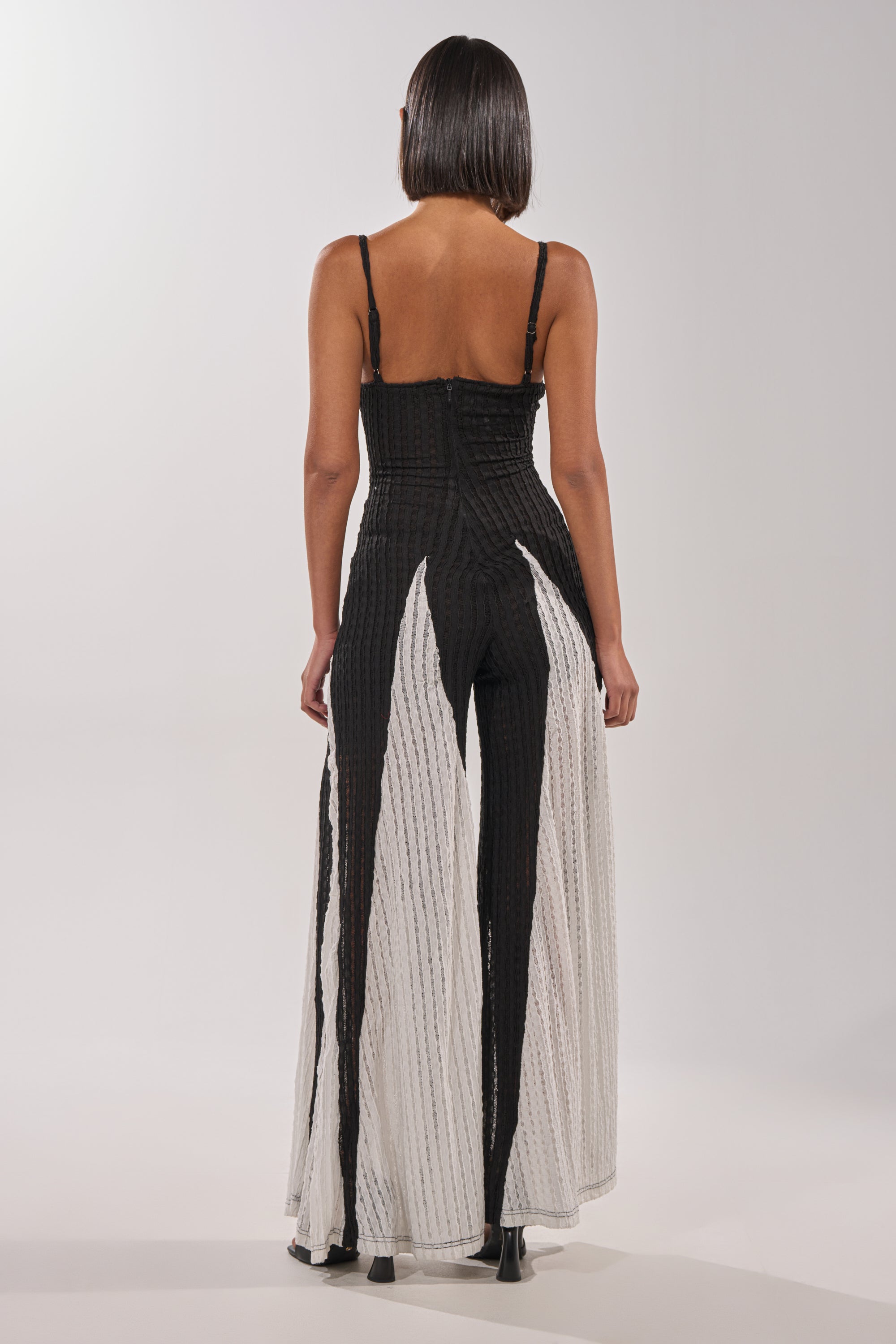 A woman with straight, shoulder-length hair faces away from the camera, wearing the ALL DAY EVERYDAY COLOR BLOCK WOVEN JUMPSUIT and strappy heels against a plain light background. 4
