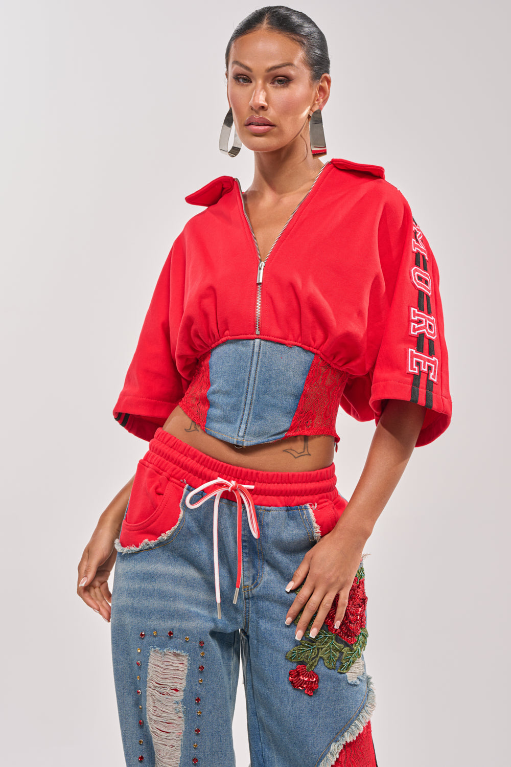 A woman wears the GAME TIME SWEATSHIRT—red, varsity style with "FIRE" on the sleeve—paired with a lace-up denim corset and denim sweatpants featuring red accents and embroidered roses, standing confidently against a light background. 0