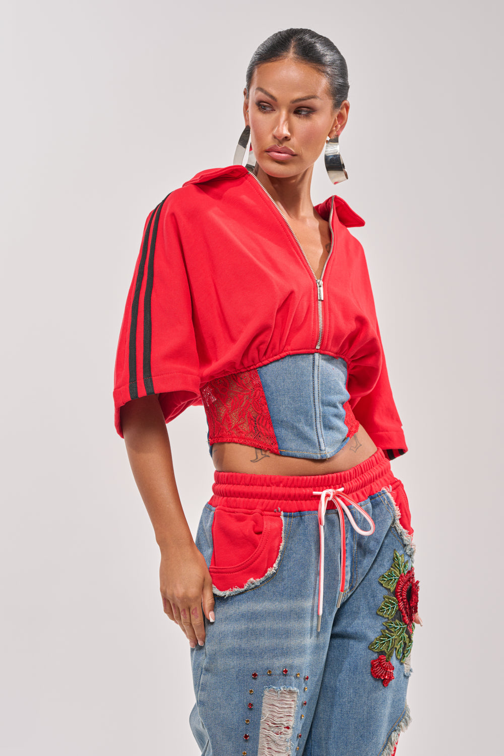 A woman models the GAME TIME SWEATSHIRT, a denim and red lace varsity corseted style with athletic stripe details, paired with blue jeans featuring red pockets and floral embroidery. She accessorizes with large hoop earrings and slicked-back hair. 1