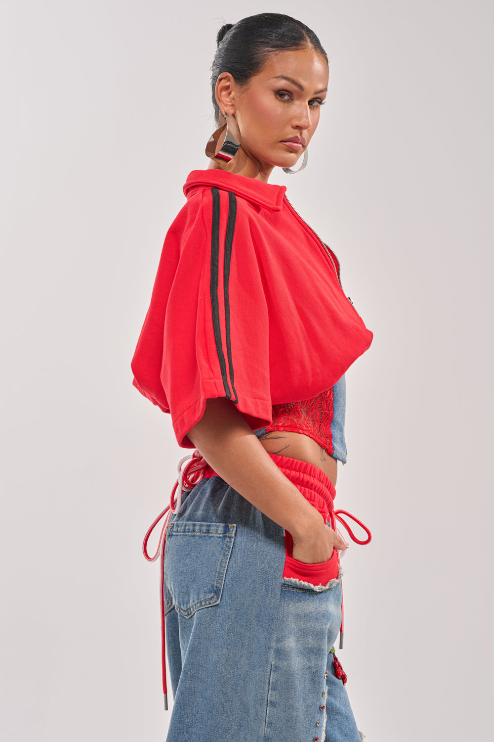 A woman with dark hair tied back models the GAME TIME SWEATSHIRT, featuring athletic-inspired sleeve stripes, paired with high-waisted blue jeans with red details. She stands in profile against a plain light background, looking at the camera. 2