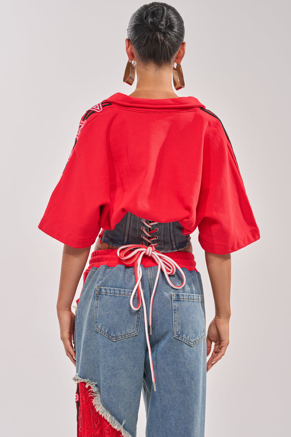 A person faces away, wearing the GAME TIME SWEATSHIRT—a red varsity corseted top with wide sleeves—paired with blue jeans featuring red fringe, a denim lace-up corset belt, bold earrings, and a neat bun. 3