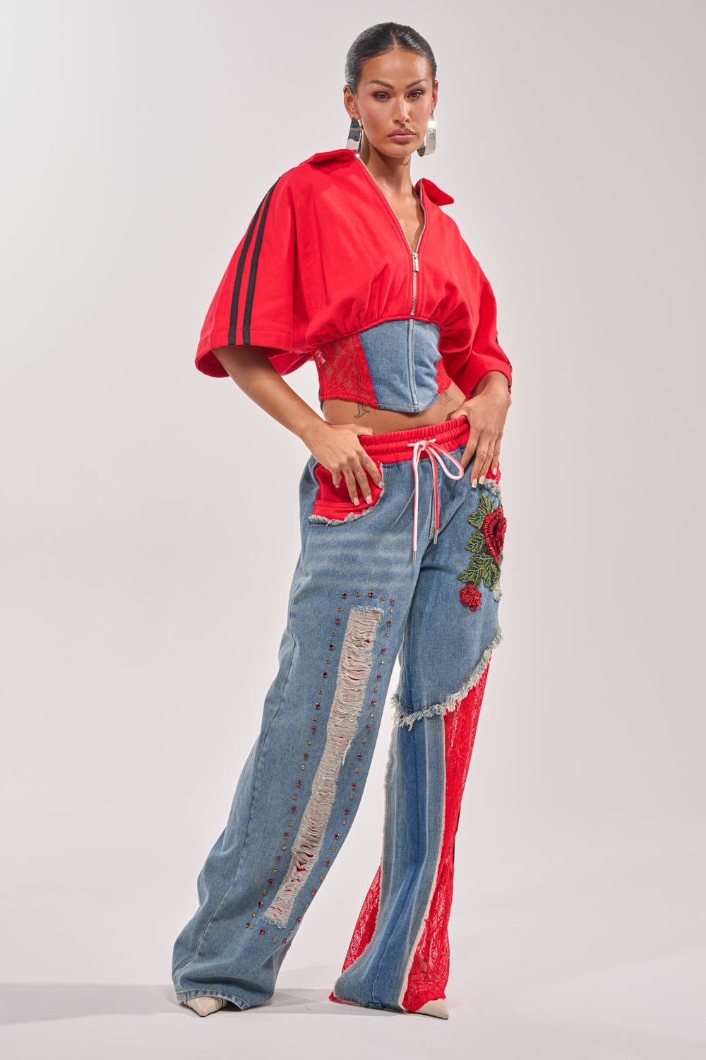 A woman poses confidently in the GAME TIME SWEATSHIRT, a varsity red corseted crop top, paired with high-waisted patchwork jeans featuring red lace, floral embroidery, and distressed details against a neutral backdrop. 5