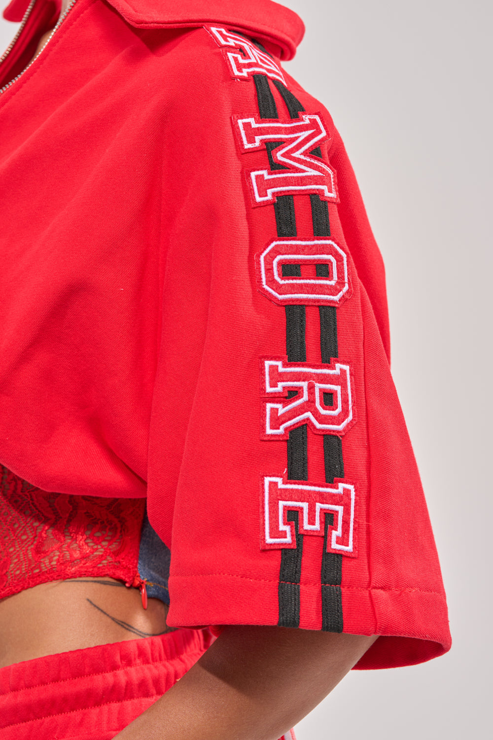 A close-up of a person wearing the GAME TIME SWEATSHIRT, varsity red with "AMORE" in bold white and red letters outlined on the sleeve beside two black stripes, set against a plain gray background. 6