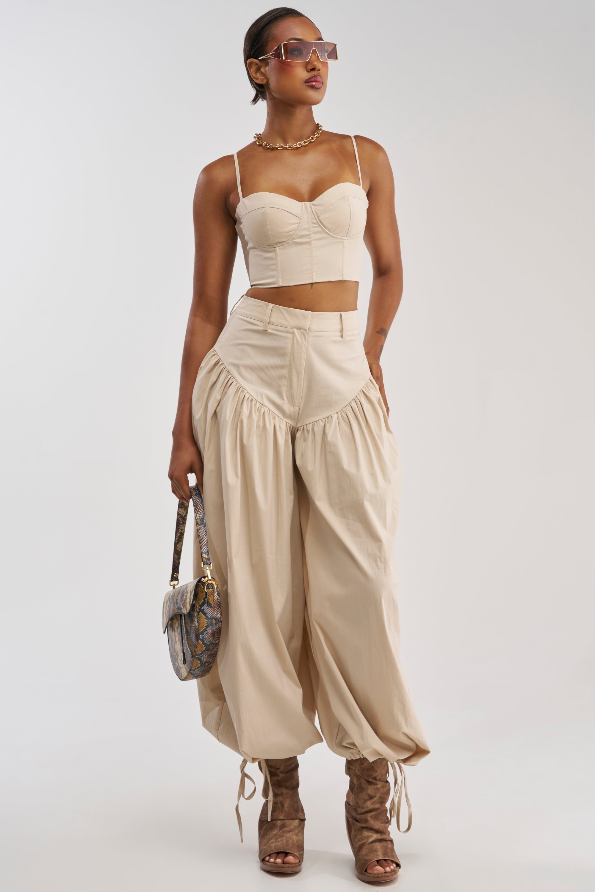A woman poses in the LUCKY FOR YOU BALLOON PANT IN BEIGE, pairing the loose, gathered pants with a beige crop top for effortless summer style. She completes her look with sunglasses, a necklace, brown heeled boots, and a patterned handbag against a plain background.