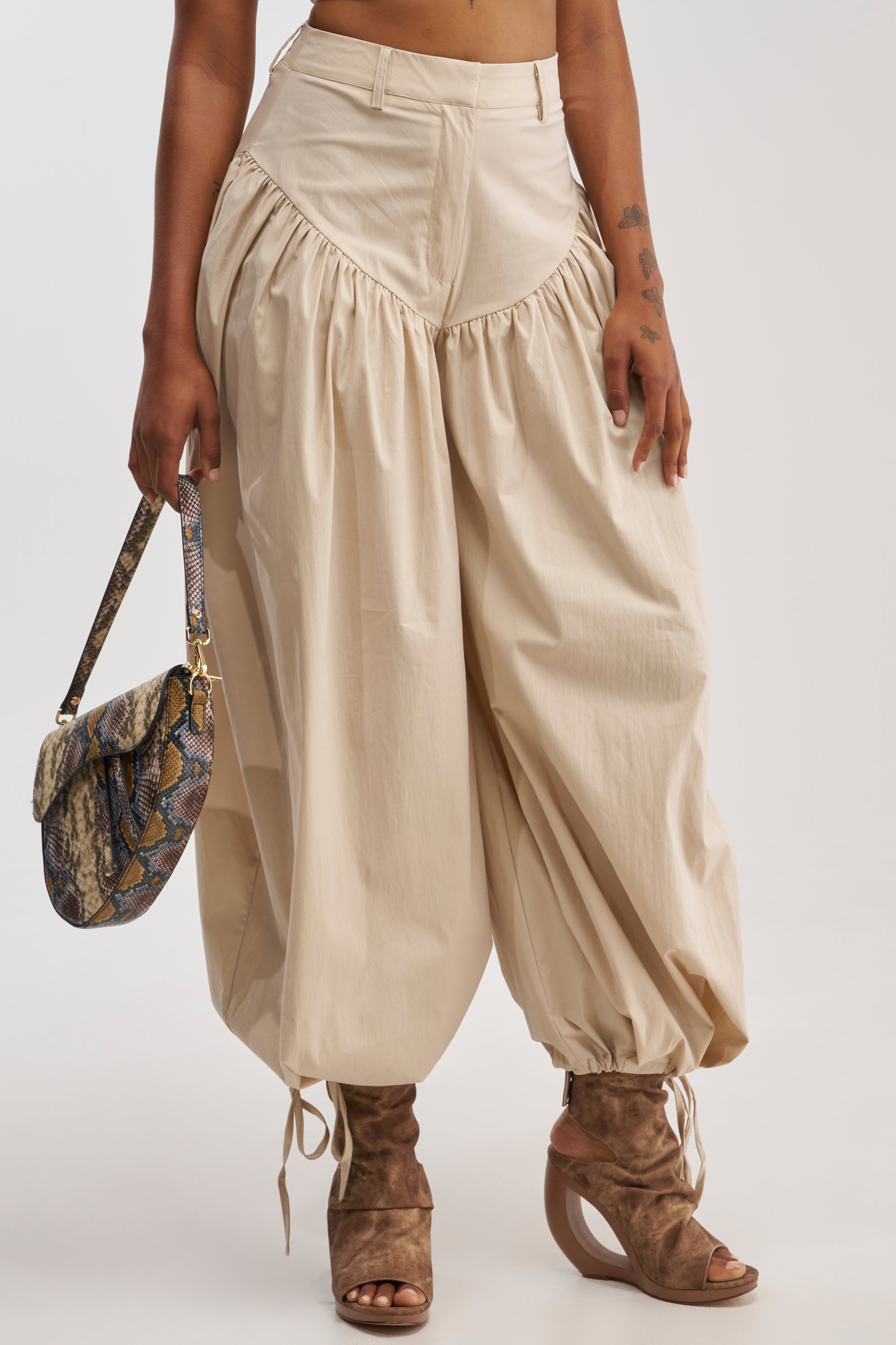A person is seen from the waist down, wearing the LUCKY FOR YOU BALLOON PANT IN BEIGE styled in a loose harem silhouette, paired with taupe open-toe wedge boots and holding a small snakeskin-patterned handbag against a plain background.