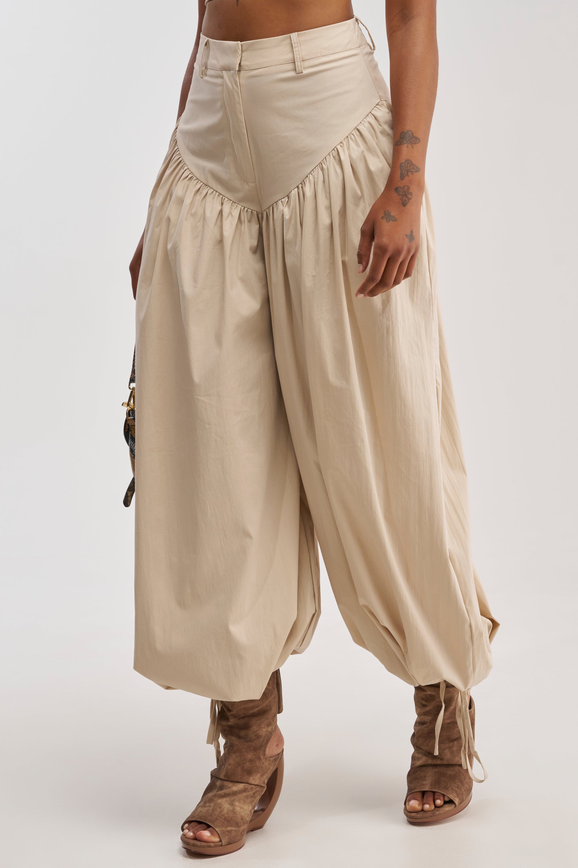 Wearing the LUCKY FOR YOU BALLOON PANT IN BEIGE, styled in a wide-legged balloon silhouette with gathered seams, this person pairs them with brown open-toe heeled boots. Their star-tattooed hand holds a small handbag, showcasing only the lower body—a perfect showcase of summer statement pants in action.