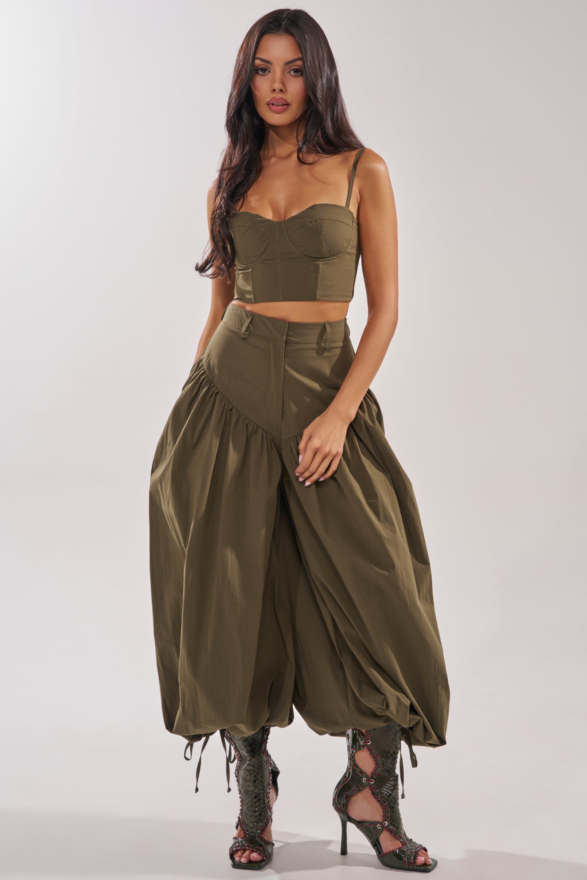 A woman with long dark hair wears an olive green bustier crop top and the LUCKY FOR YOU BALLOON PANT, styled with lace-up peep-toe boots. She stands confidently against a plain, light background. 0