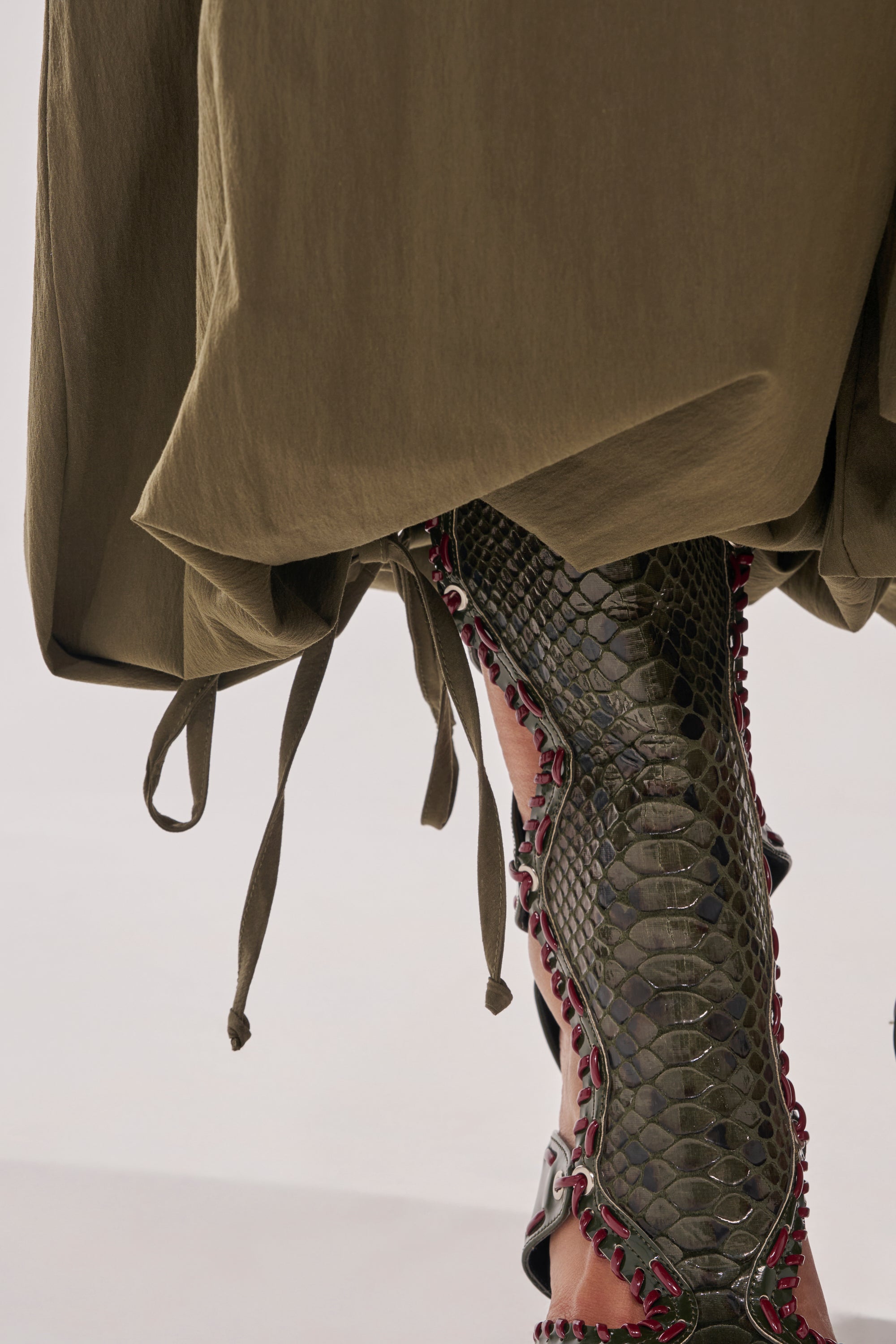 A close-up of a person wearing the LUCKY FOR YOU BALLOON PANT in olive green, styled with dark green snakeskin knee-high boots featuring red studded accents. 9